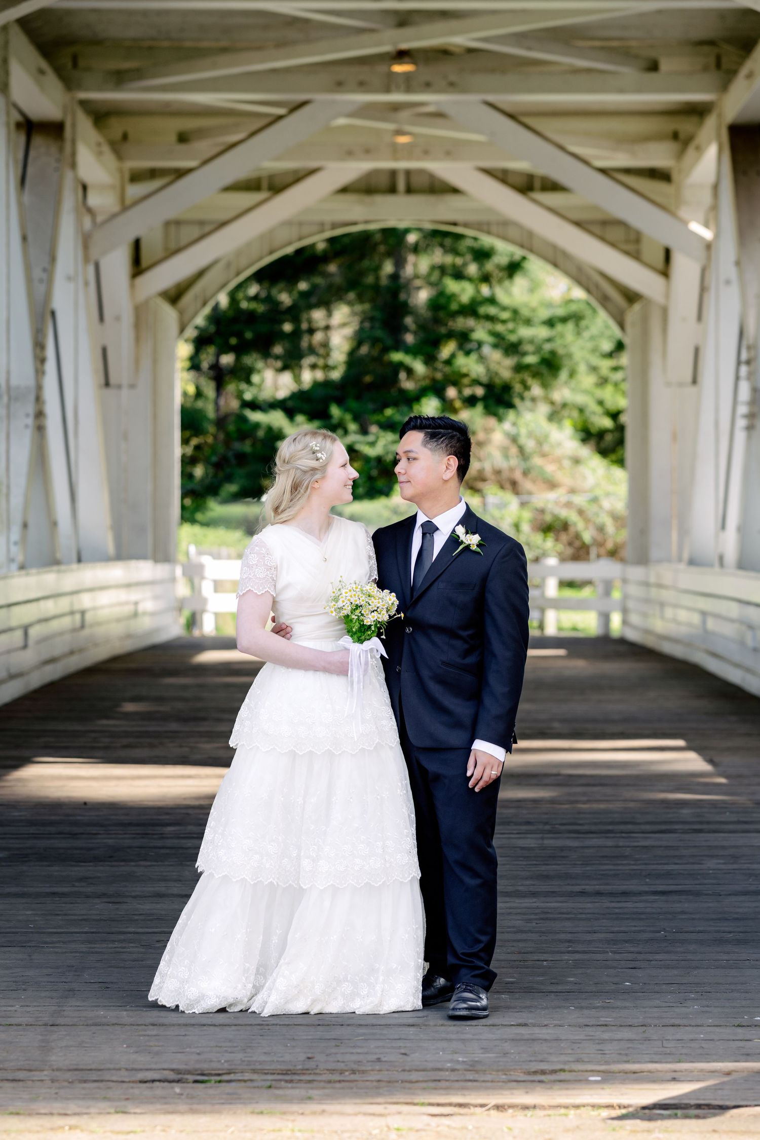 A bride and groom share a romantic moment on a white covered bridge surrounded by lush greenery.