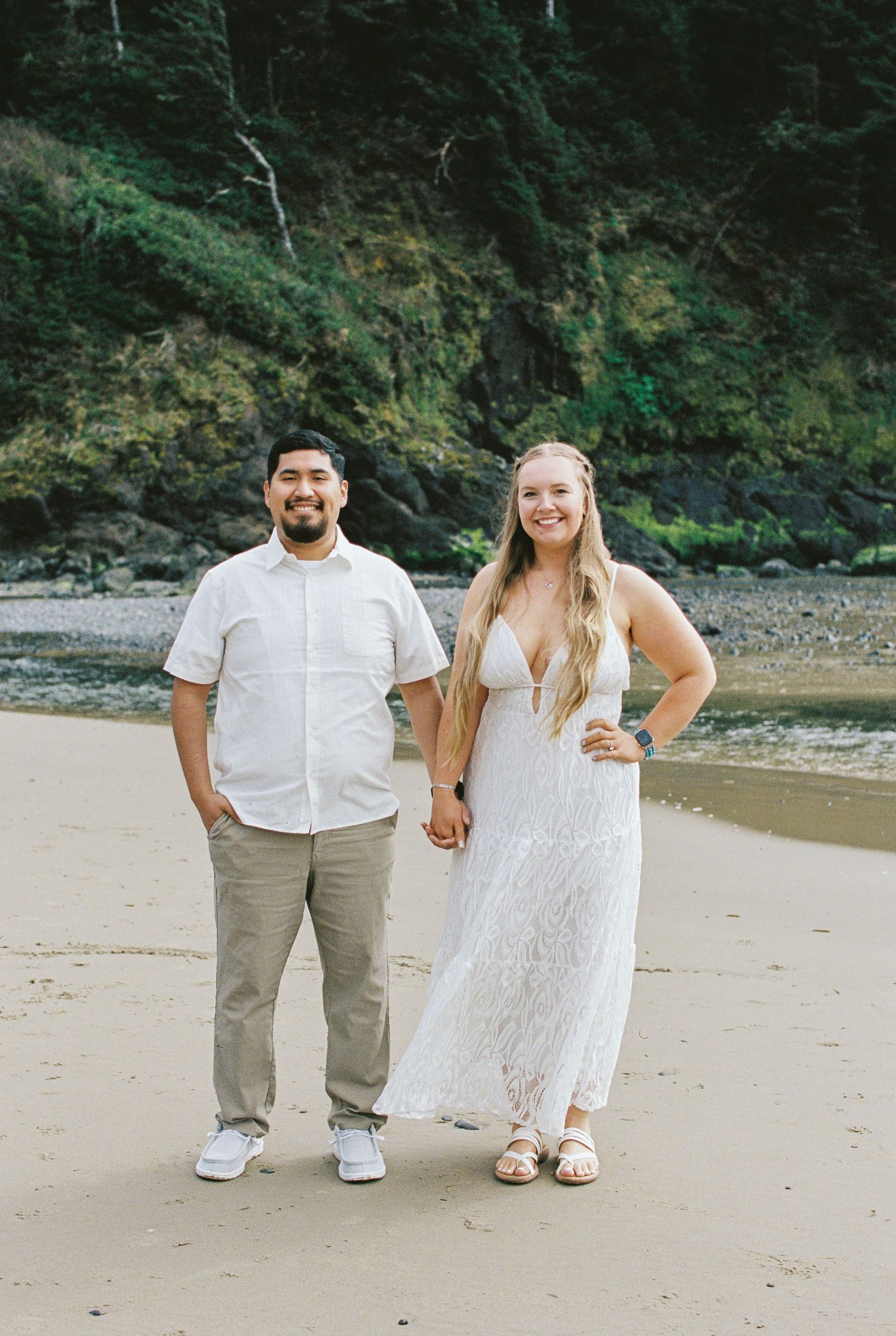 Smiling couple holding hands in white outfits on Oregon coast beach with forest backdrop.
