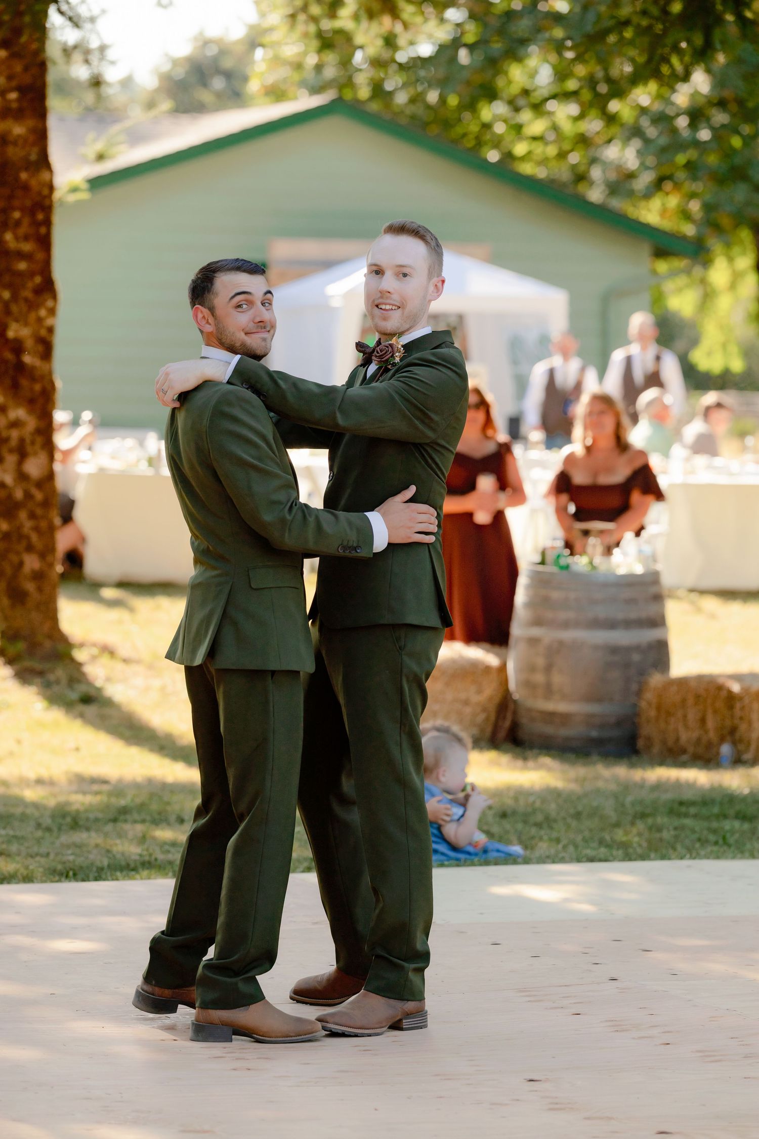 Two grooms in matching olive green suits share a dance together at their outdoor rustic wedding reception.