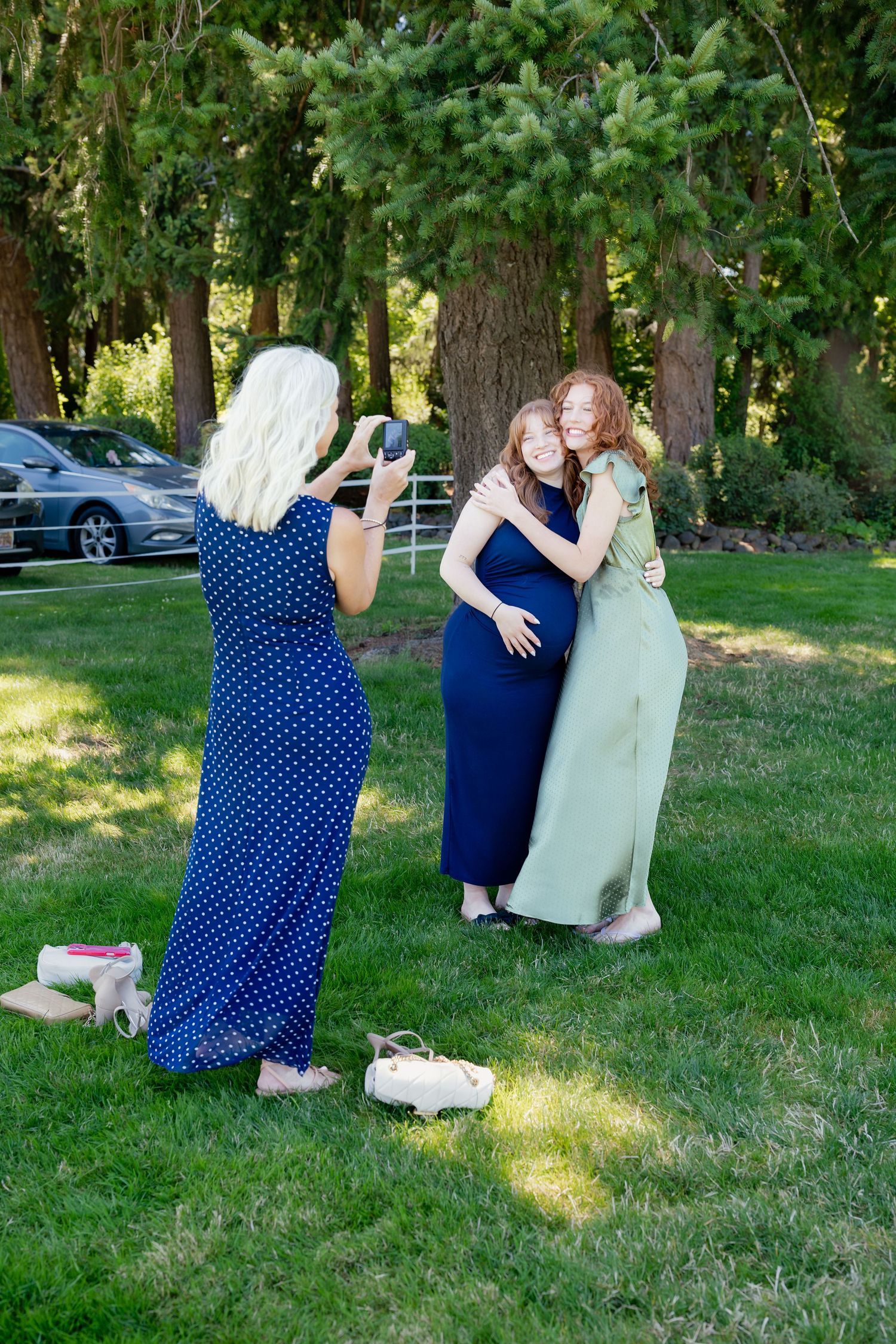 Woman in blue polka dot dress photographing two women hugging in green and navy dresses at outdoor wedding.