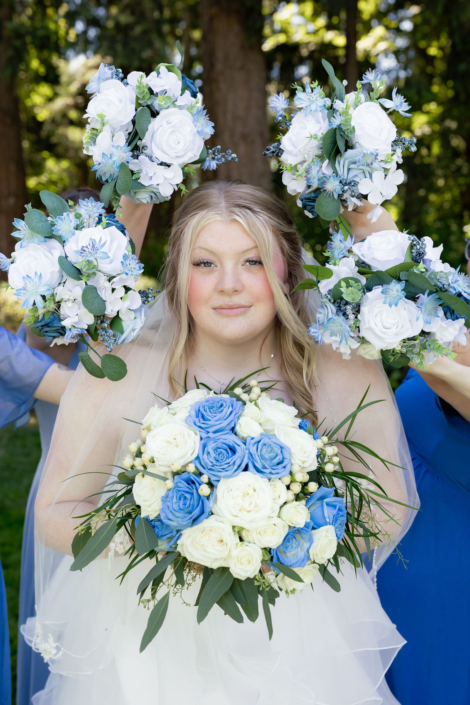 Smiling bride holding blue and white rose bouquet surrounded by bridesmaids raising matching bouquets in outdoor setting.