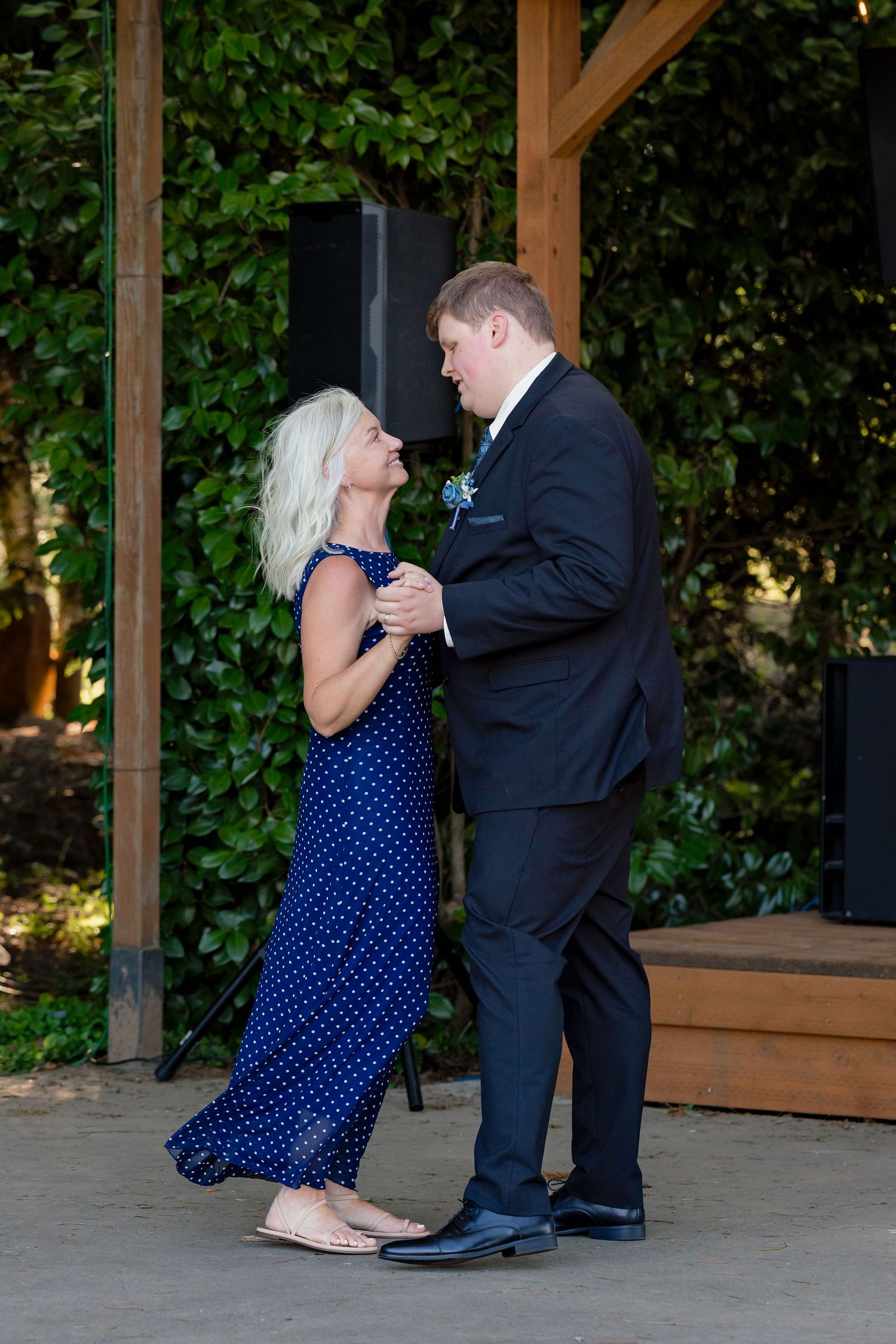 Groom in black suit dancing with woman in blue polka dot dress at an outdoor wedding reception.