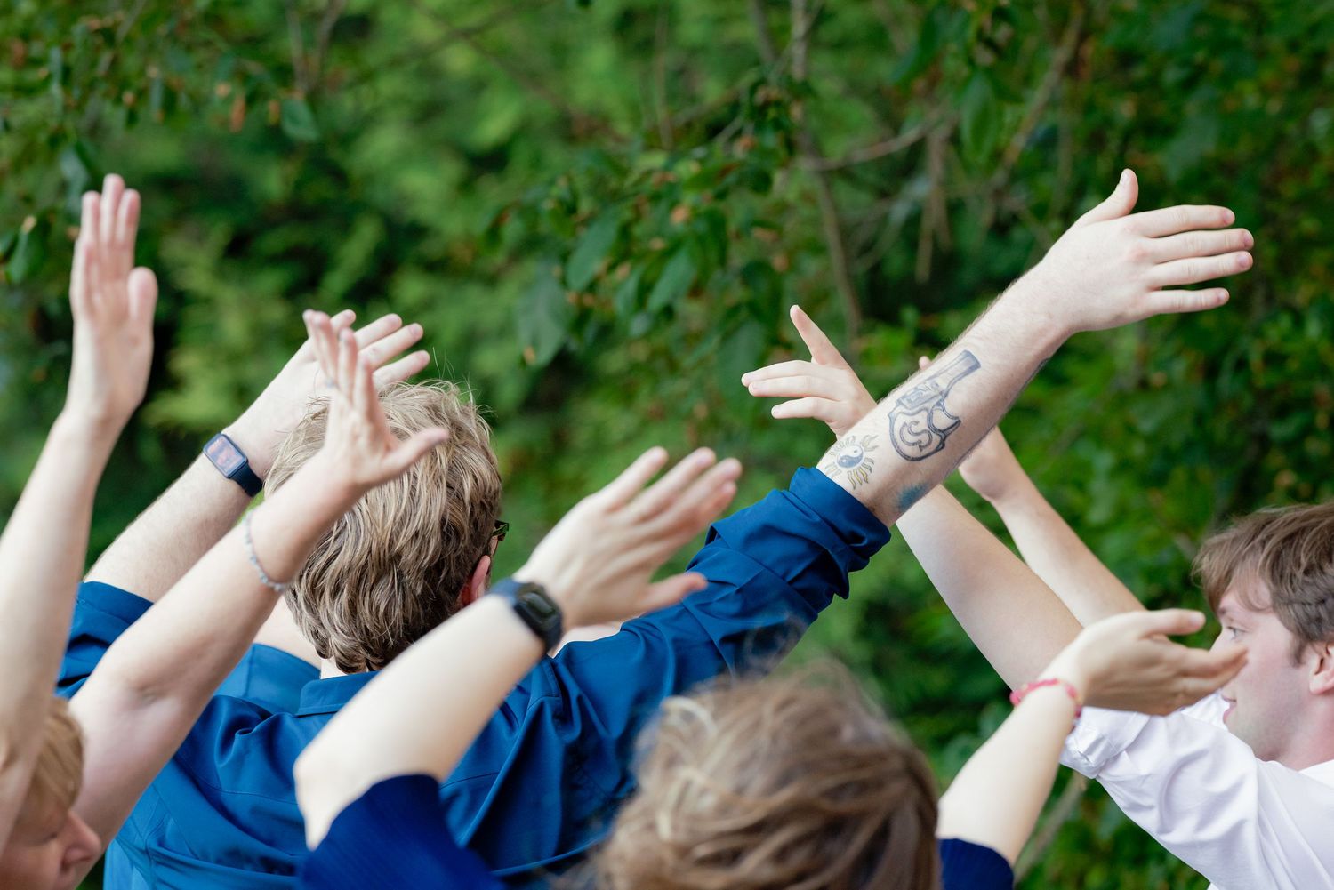 Excited wedding guests raise hands and cheer outdoors surrounded by lush green trees.
