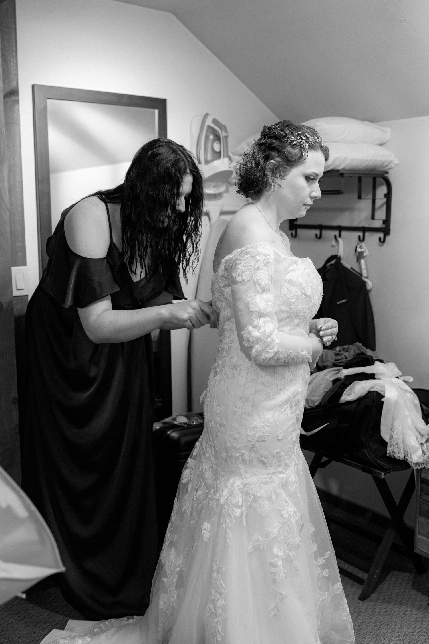Bridesmaid in black gown helping bride in lace wedding dress prepare in black and white photo.