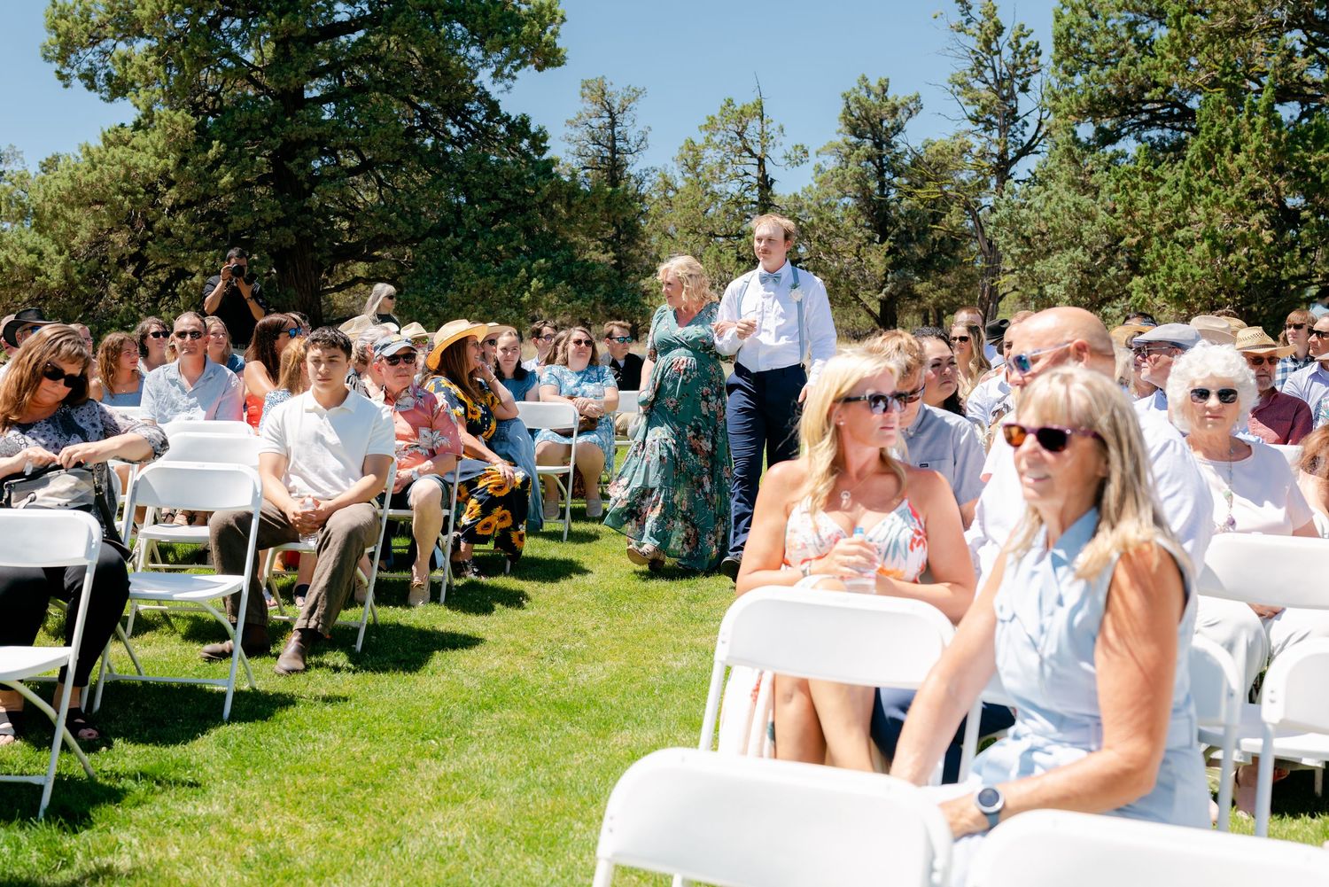 Wedding guests seated in white chairs outdoors on a sunny day, watching a ceremony on a green lawn.