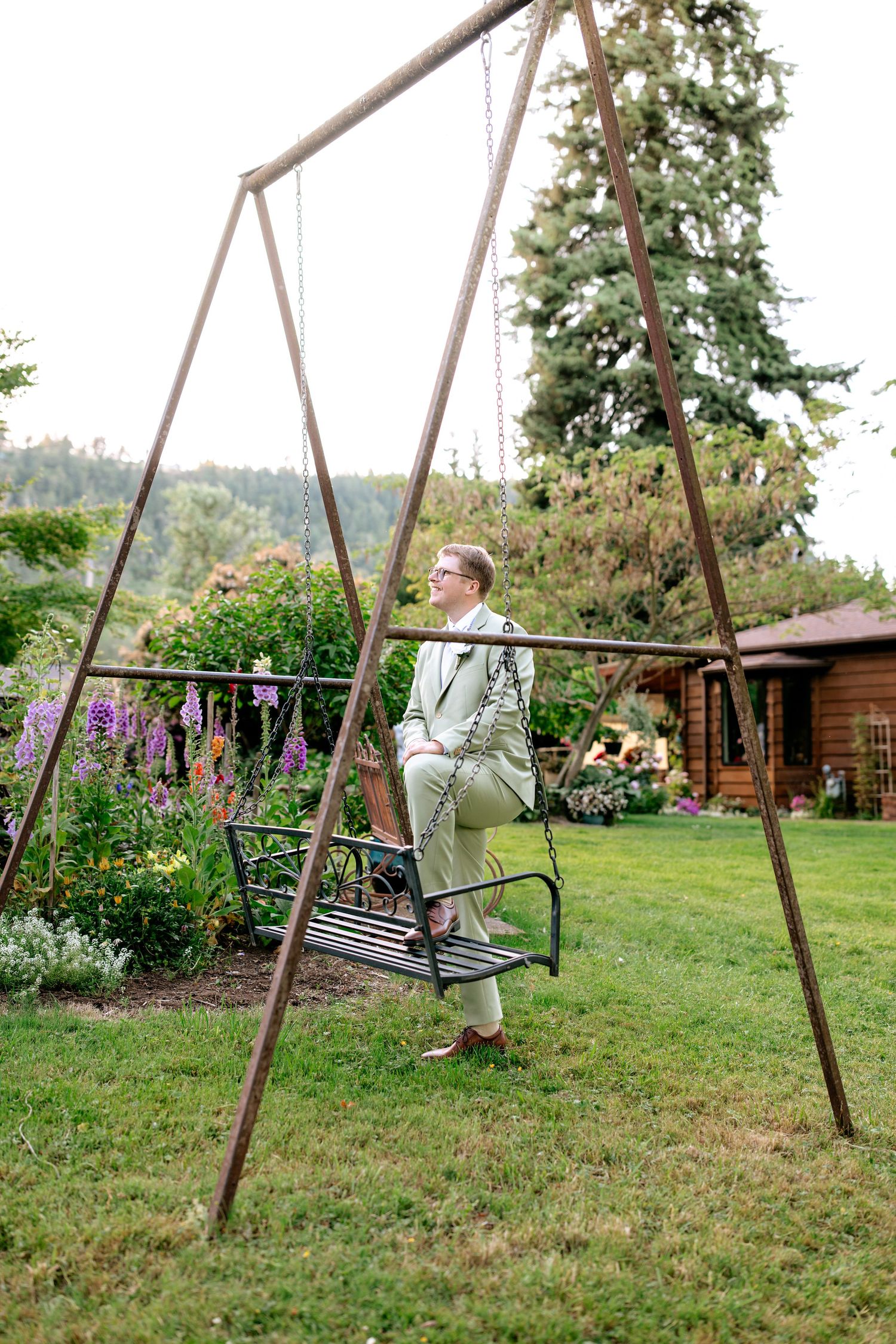 Groom in sage suit sits alone on a garden swing set surrounded by lush greenery.