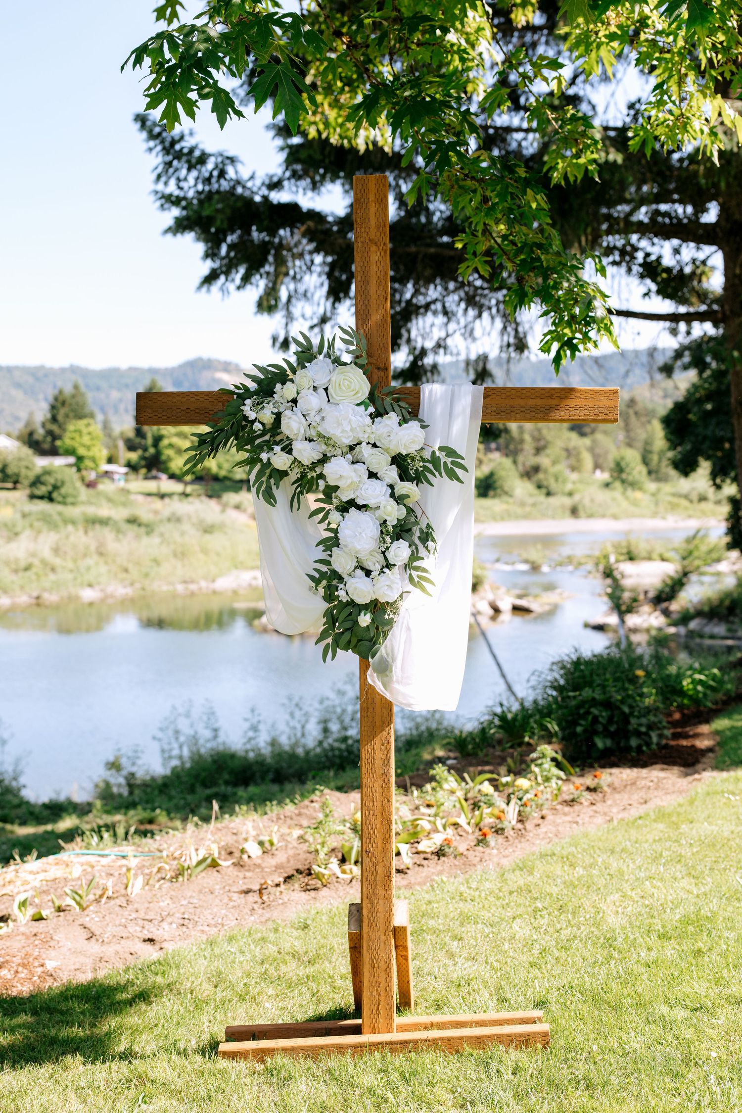 Wooden cross decorated with white flowers and greenery standing on a sunny lawn with a serene river and hills in the background.