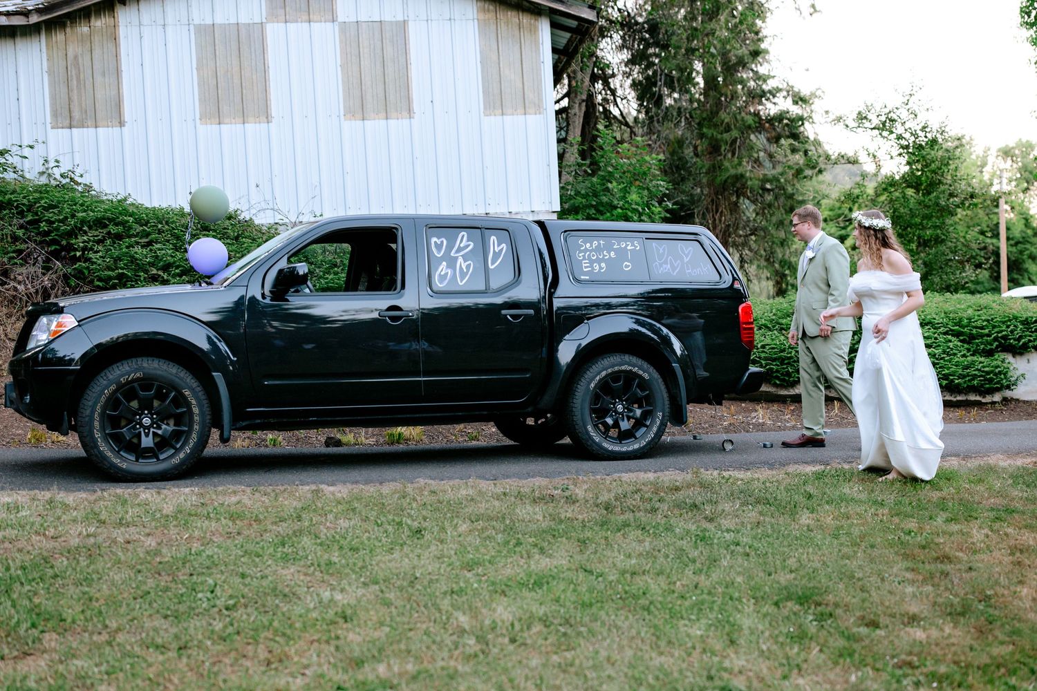 Newlywed couple stands beside decorated black pickup truck with just married signs at outdoor wedding venue.