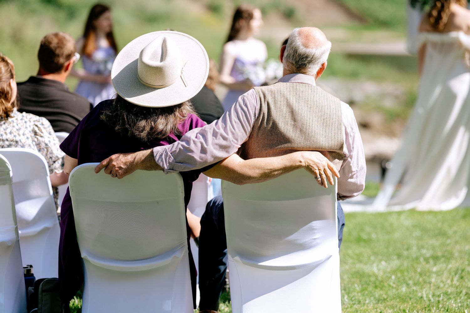Elderly couple sitting together arm in arm at an outdoor wedding ceremony on a sunny day.