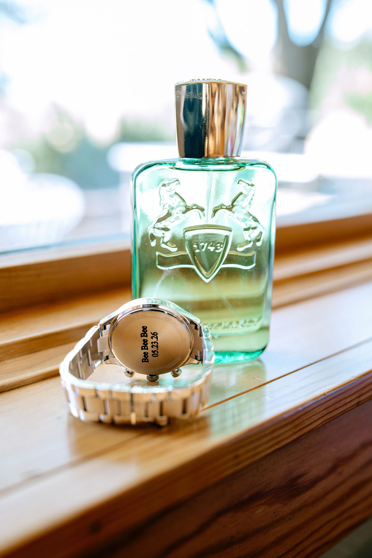 Elegant green glass perfume bottle with silver cap beside a silver watch on a wooden surface.