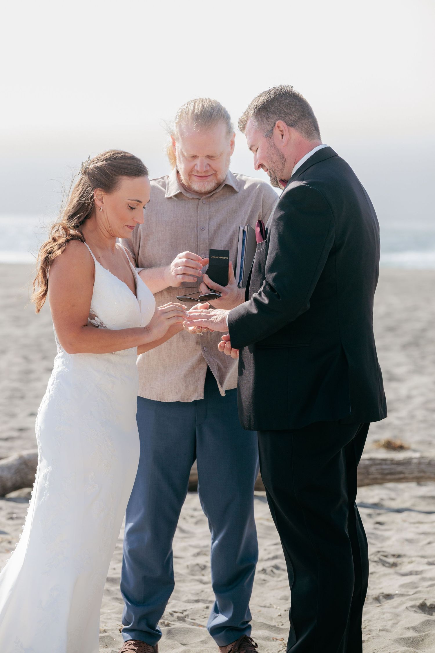Beach wedding ceremony with three people exchanging rings on a misty shoreline.