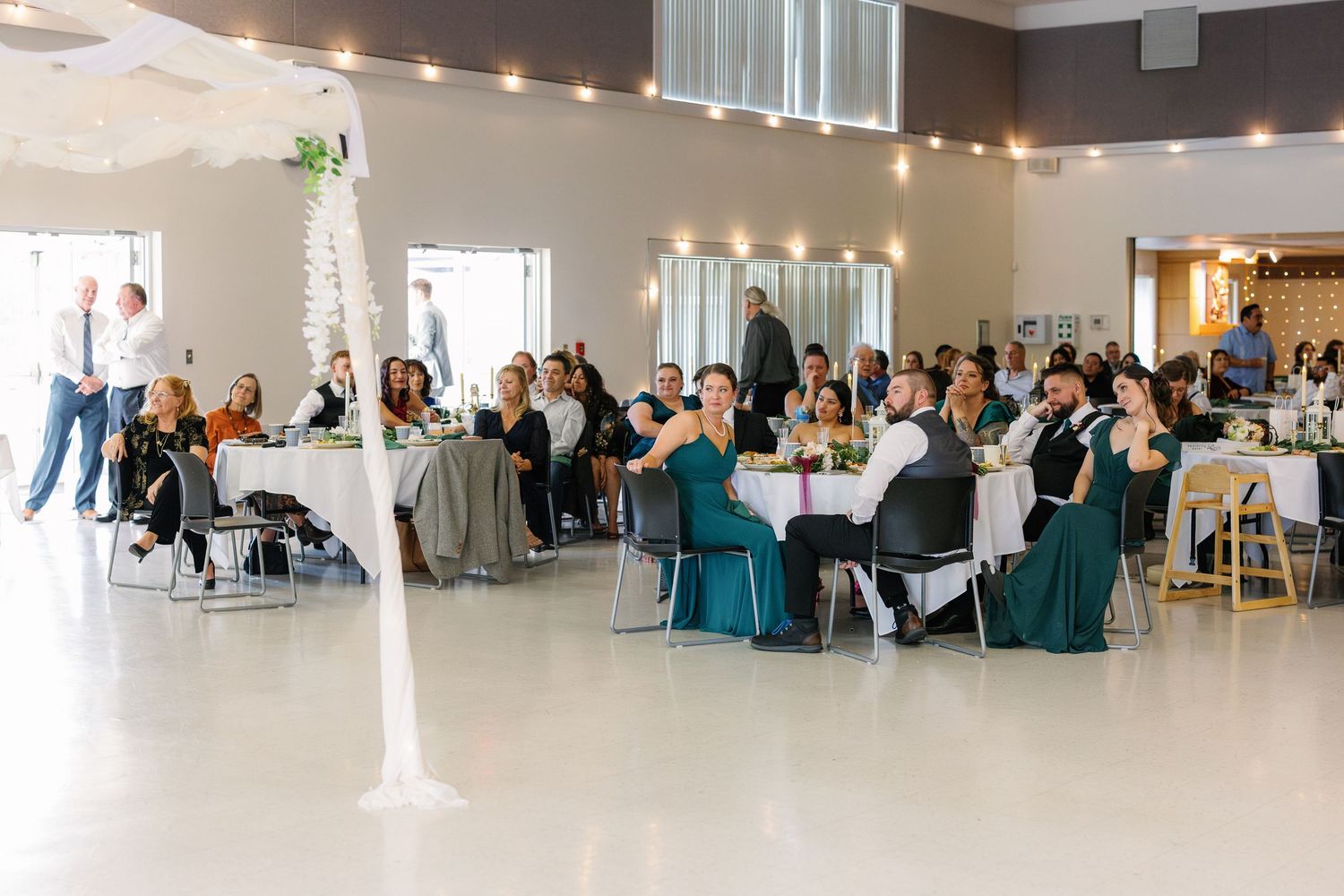 A large bright reception hall decorated with string lights and filled with wedding guests.