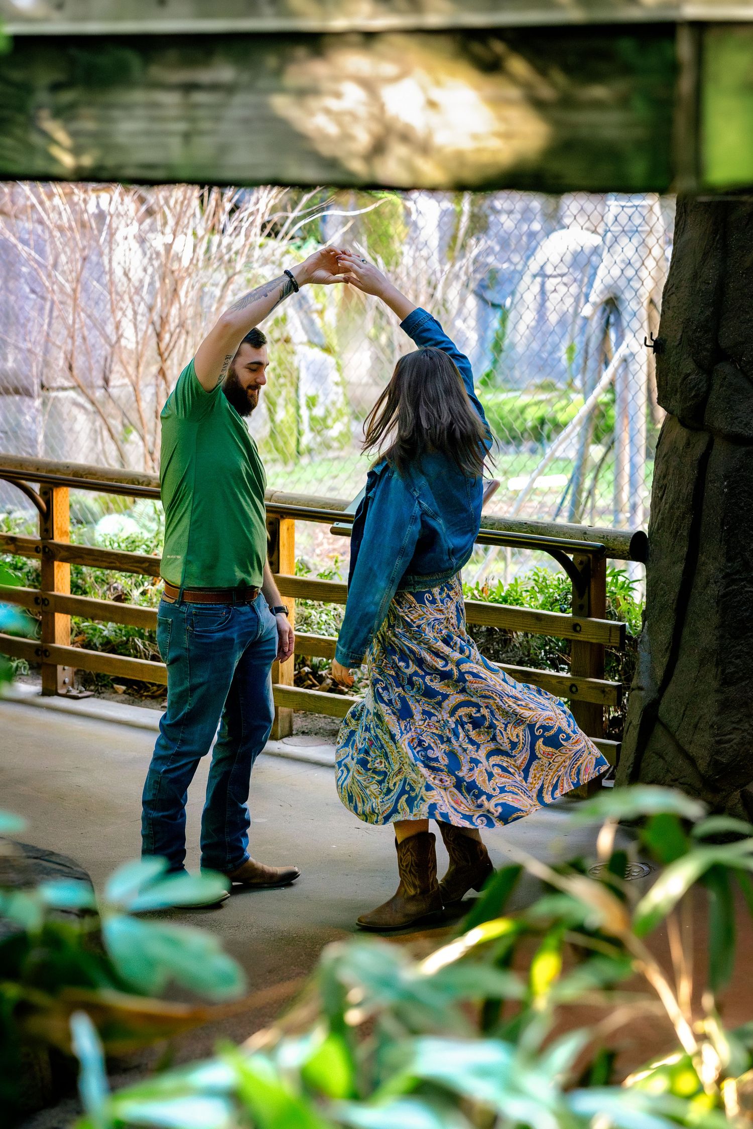 Two people in casual attire dance together on a covered walkway surrounded by greenery.
