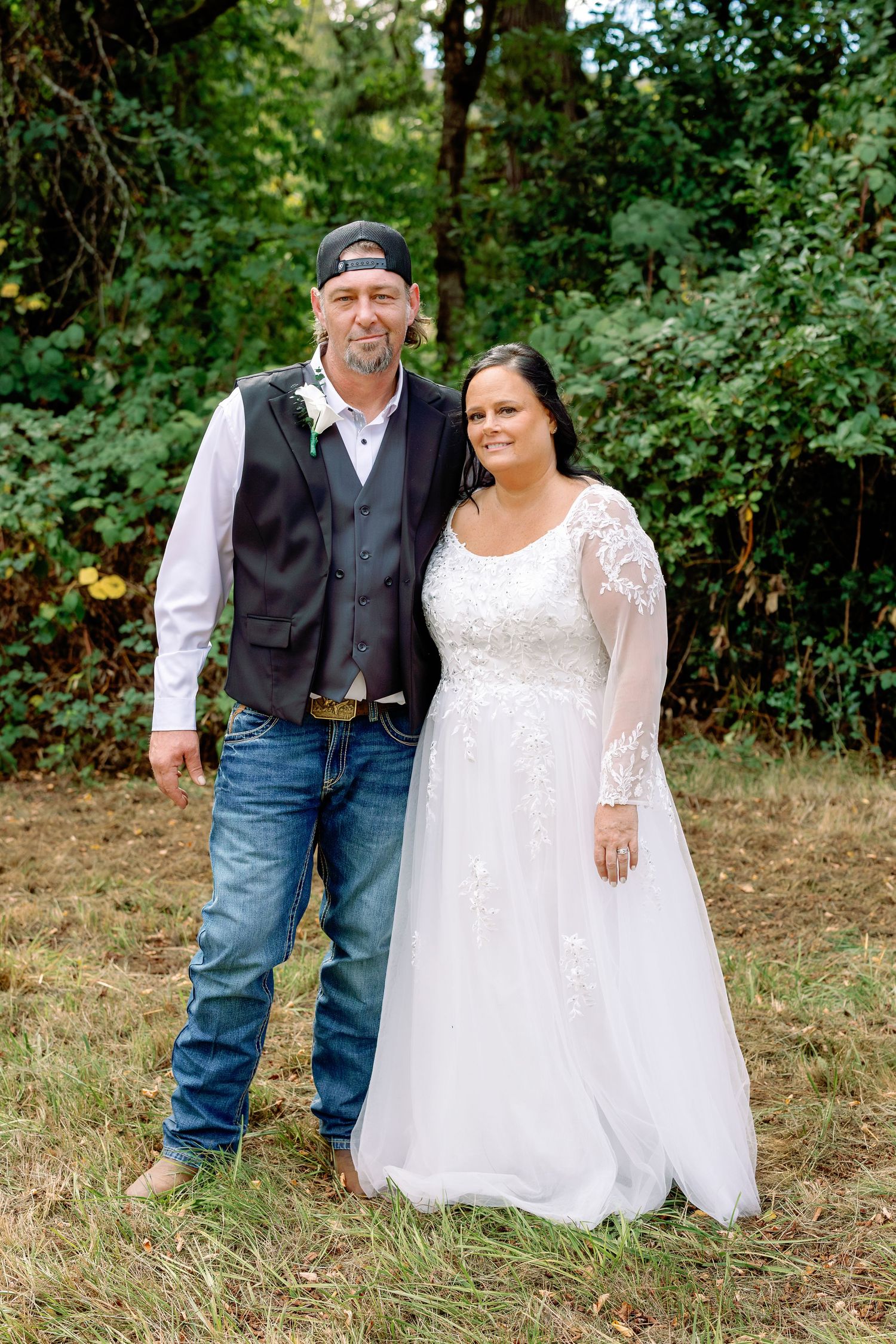A couple poses outdoors in wedding attire with the bride wearing a white lace dress and groom in casual western wear.