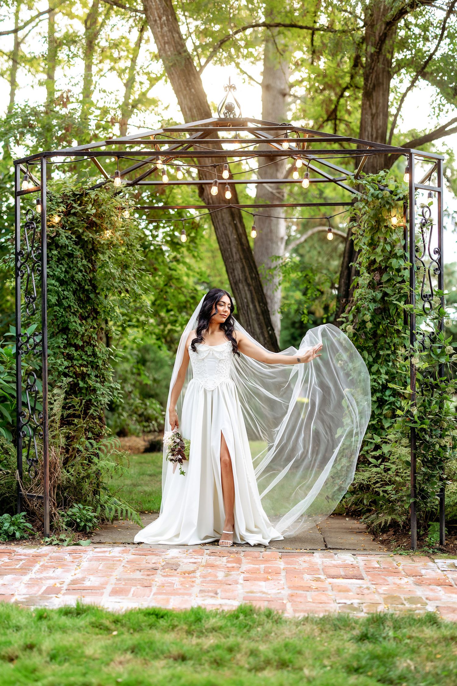 An elegant bride poses under a garden arbor with flowing white dress and veil catching the breeze.