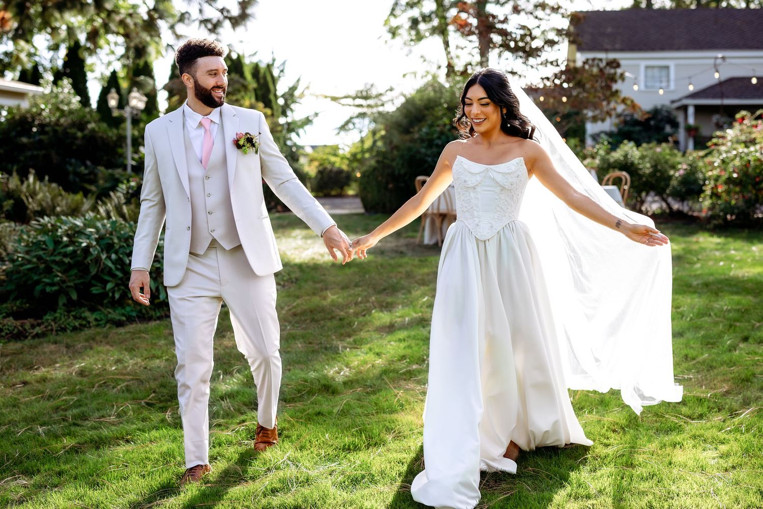 Newlyweds in white attire dance and twirl playfully on a sunlit grassy lawn.