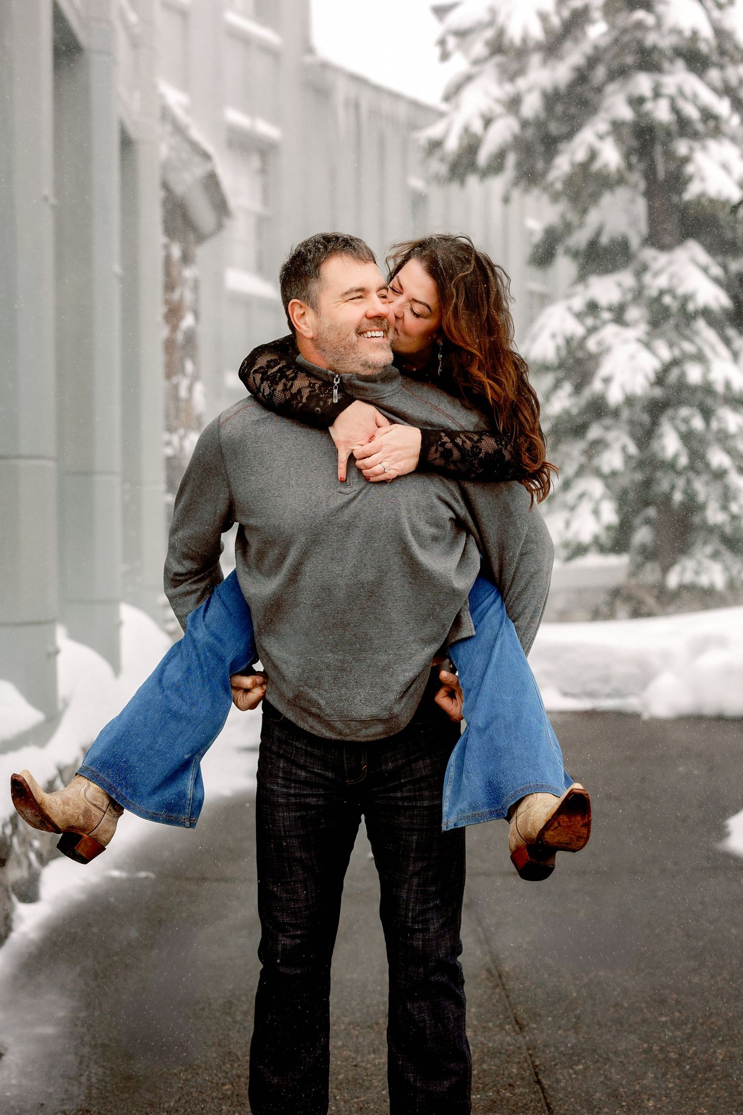 A couple shares a playful romantic moment in a snowy winter setting with evergreen trees in the background.