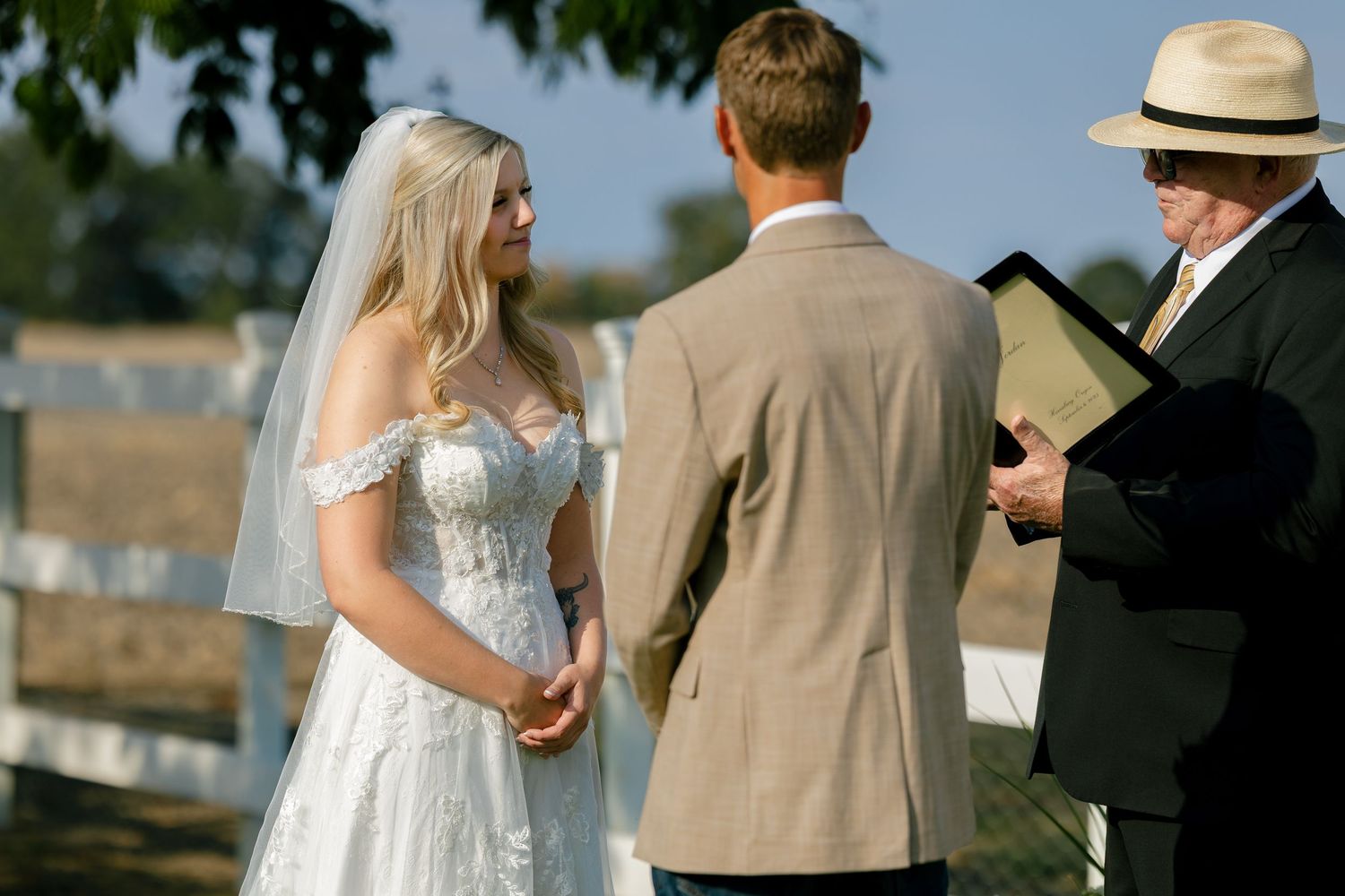 Outdoor wedding ceremony with officiant reading from book as couple stands before wooden fence.