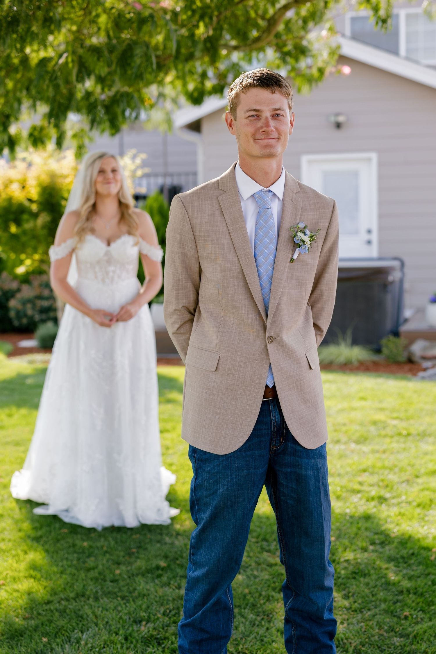 First look wedding photos in sunlit backyard with bride approaching groom in tan suit.