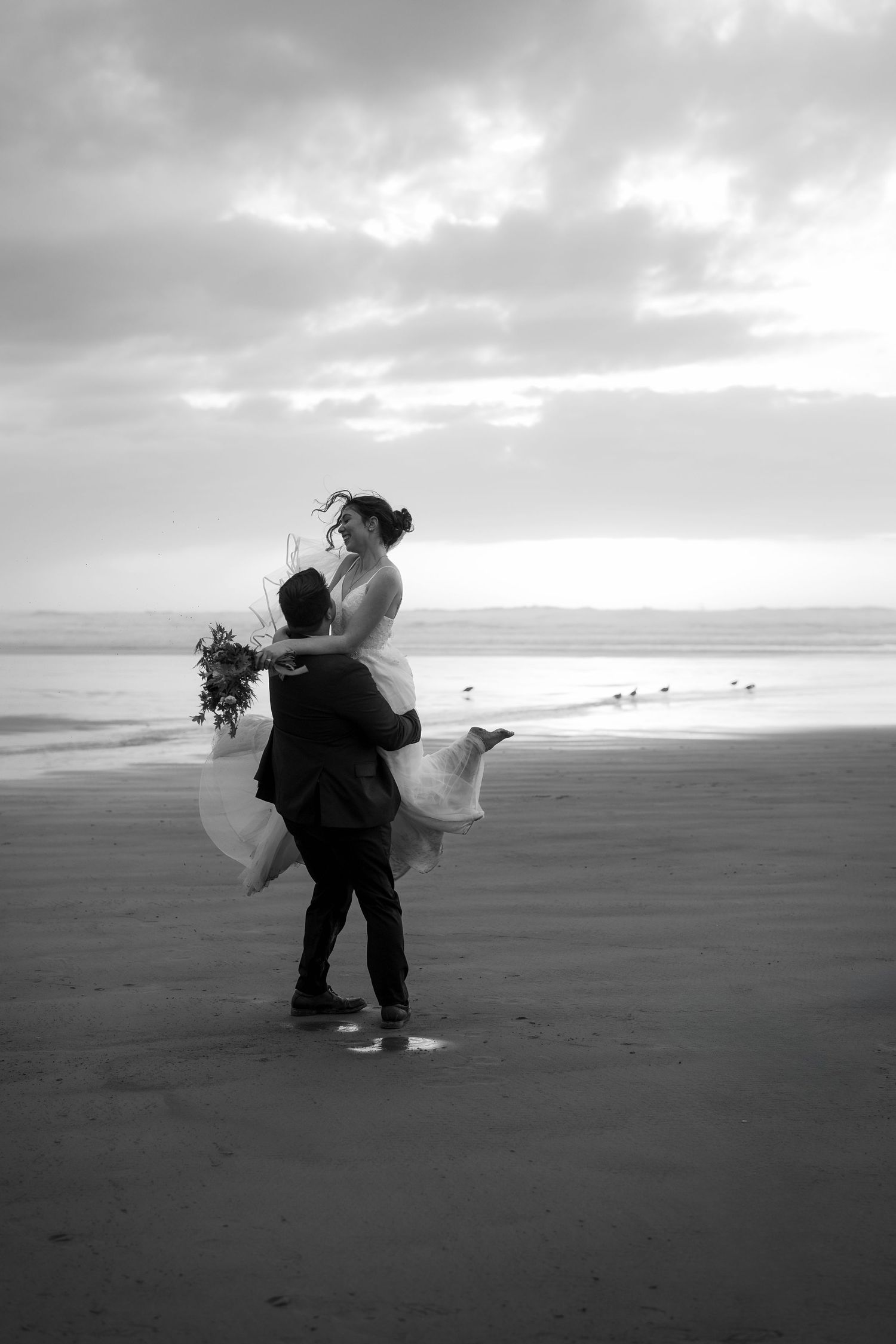 A black and white photo captures newlyweds in an intimate moment on a windswept beach.
