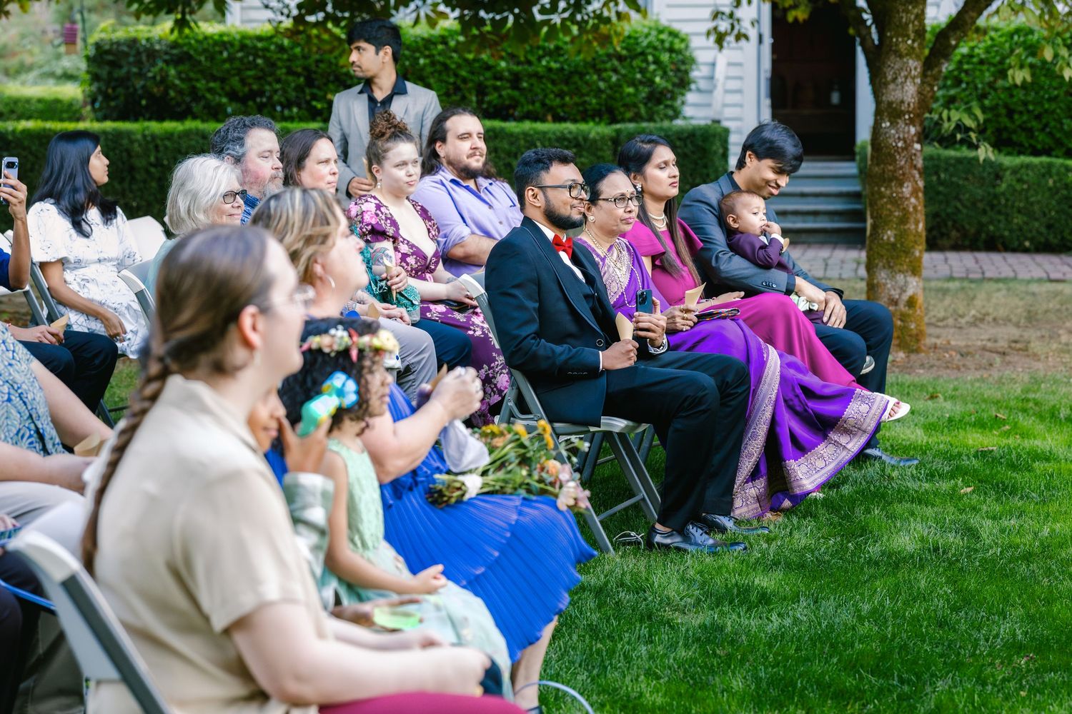Wedding guests seated on lawn chairs enjoying an outdoor ceremony in a garden setting.