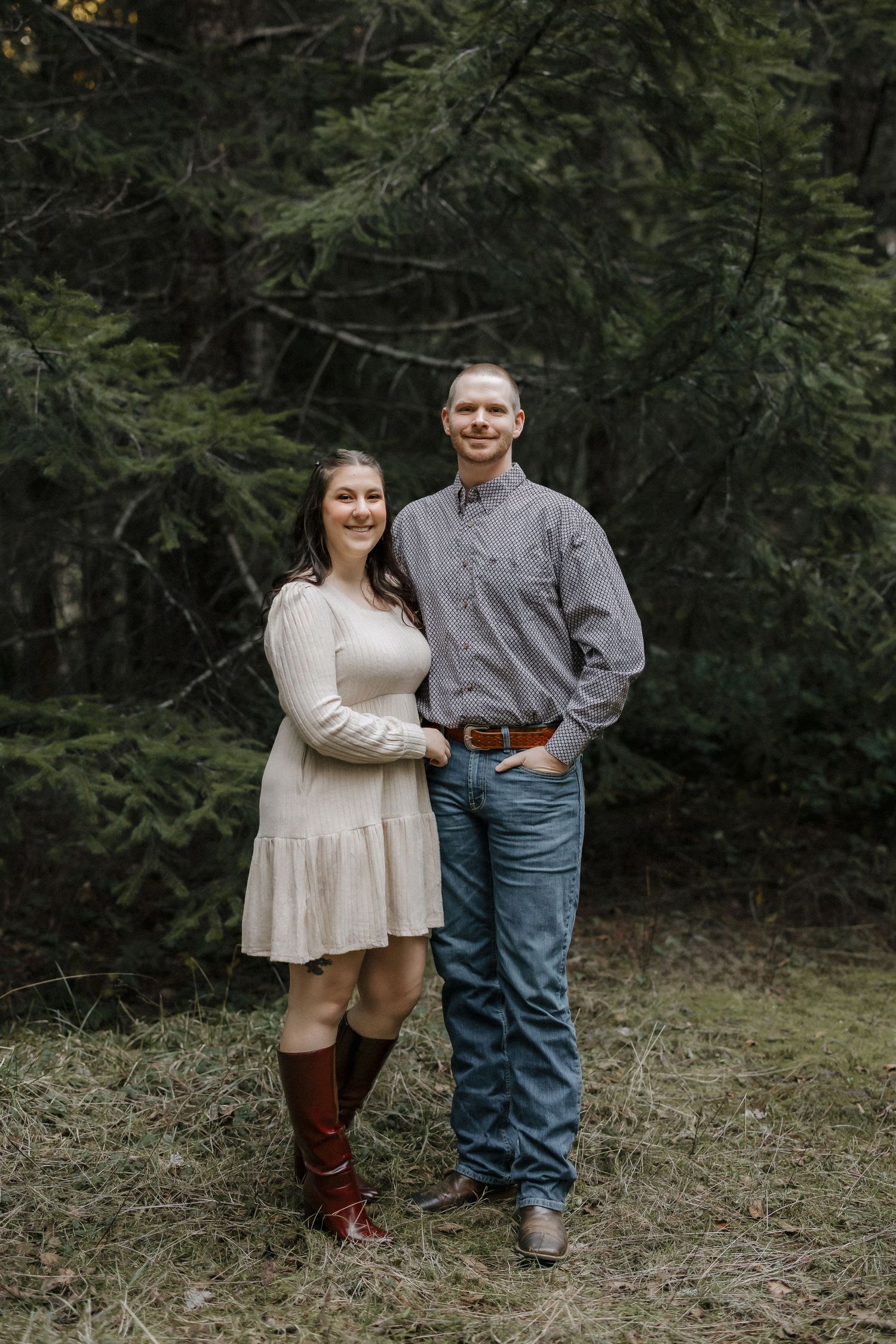 A romantic forest photoshoot featuring a couple in coordinated casual outfits against evergreen trees.