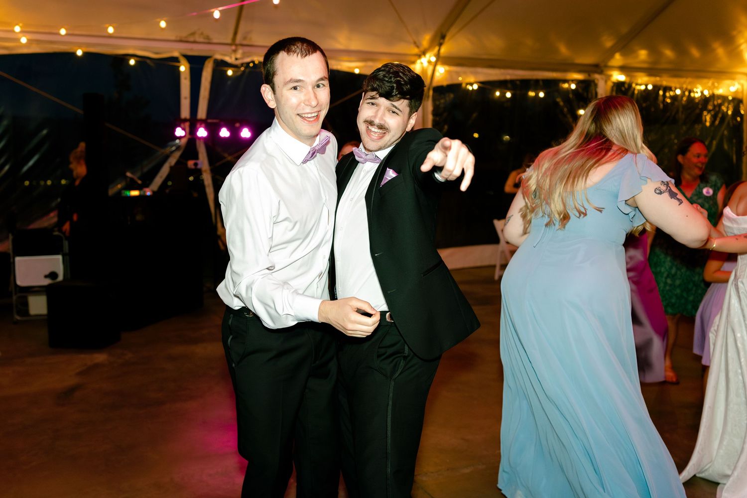 People dancing and celebrating at an evening wedding reception under a tent with string lights overhead.