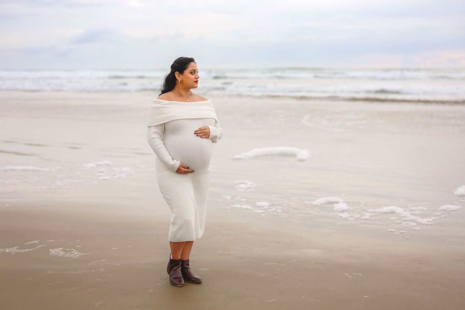 A person in a white dress walks along a misty beach shoreline during an overcast day.