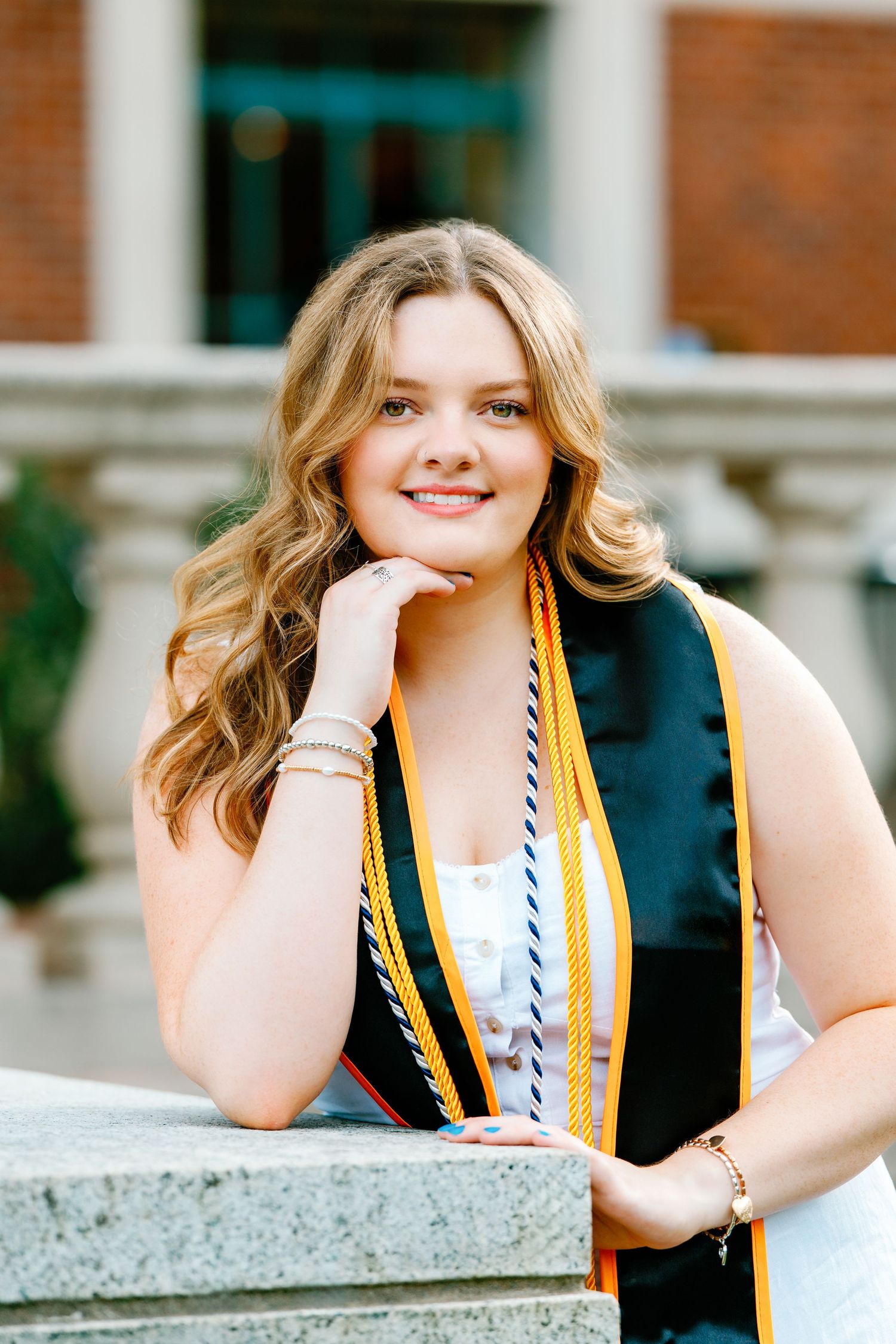 Smiling graduate poses in black and gold graduation stole against brick building backdrop.