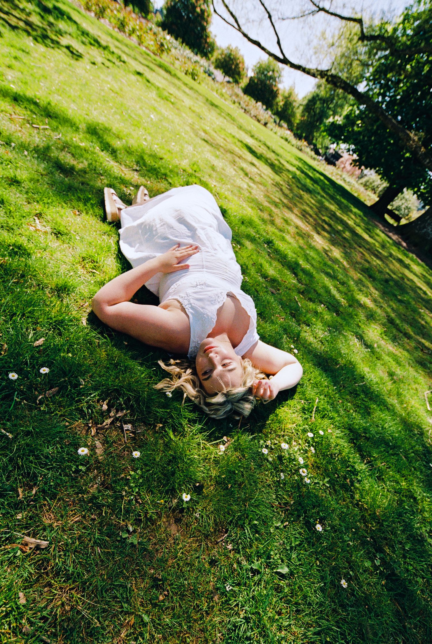 Blonde woman in white dress lying on lush green grass in sunny park setting.