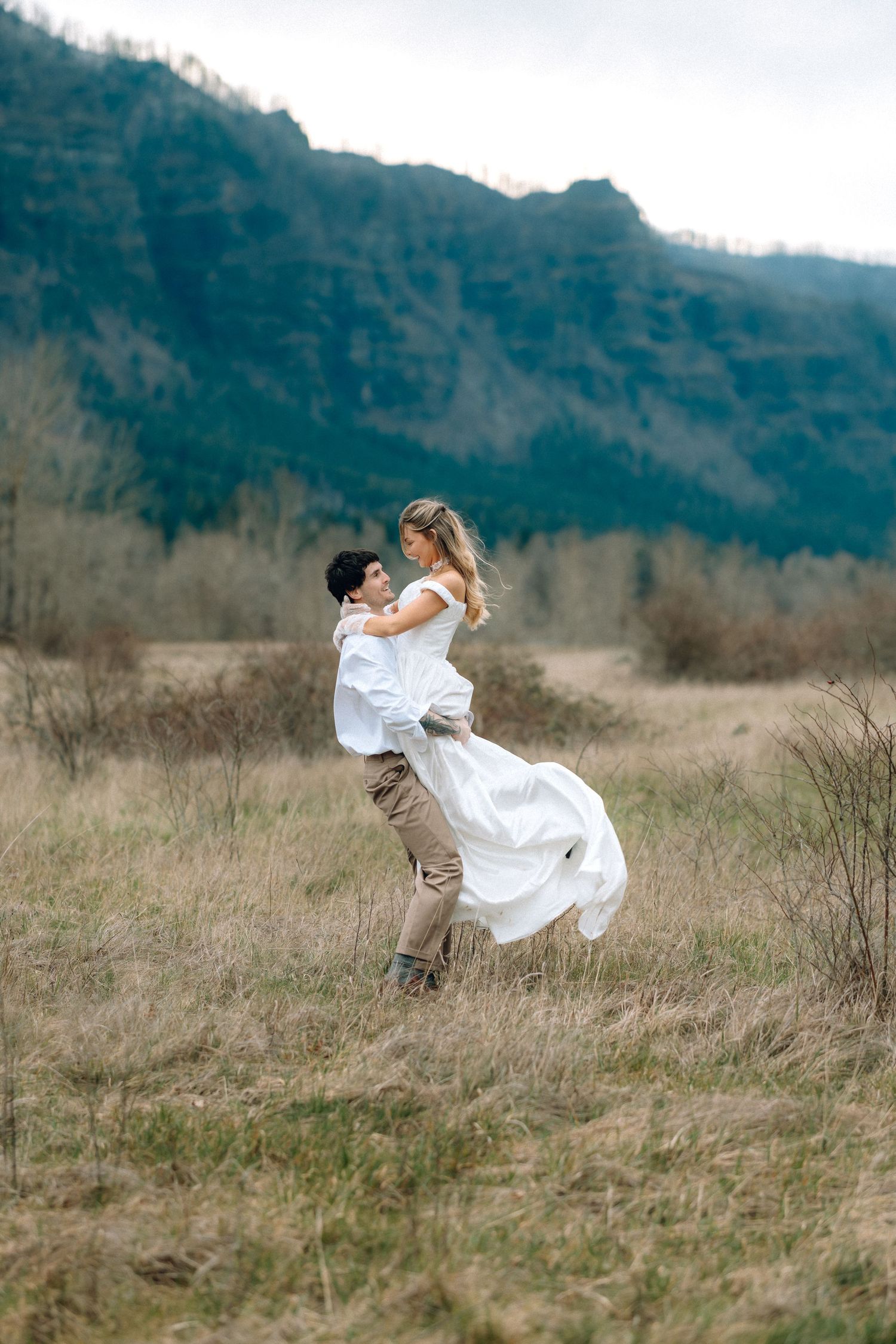 A romantic couple dances and spins in a grassy field with mountains in the background.