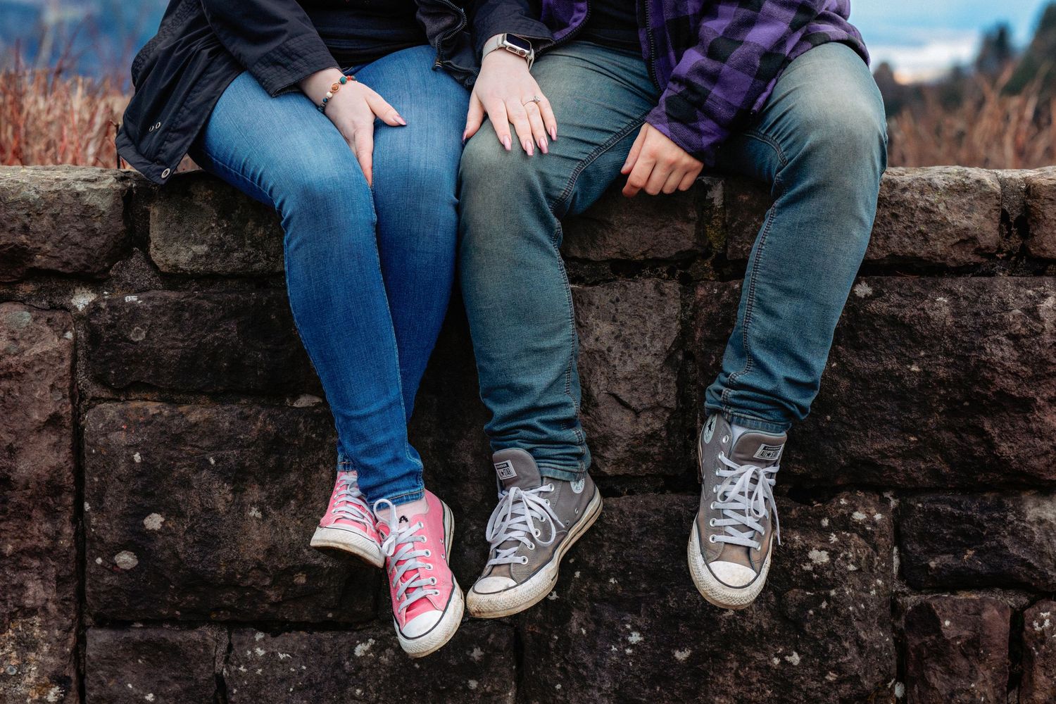 A row of feet wearing Converse sneakers and jeans lined up against a rustic brick wall.