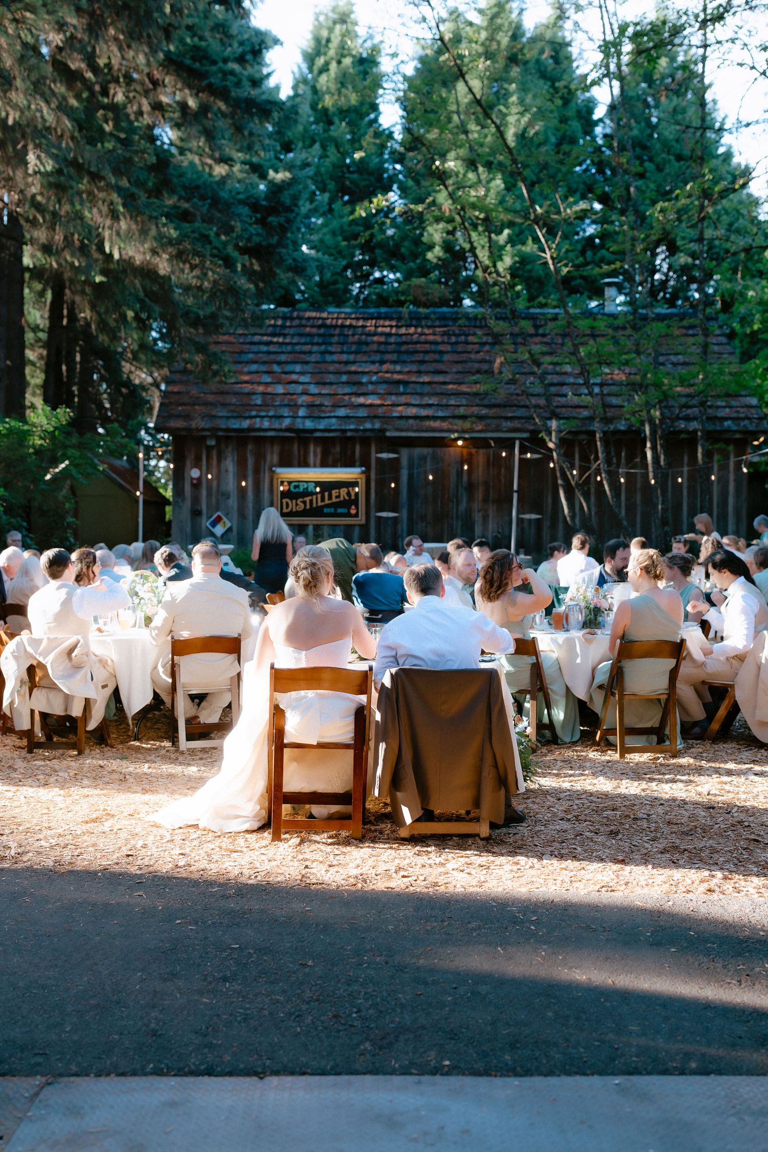 Outdoor rustic barn wedding reception with guests seated at white-clothed tables under tall trees.