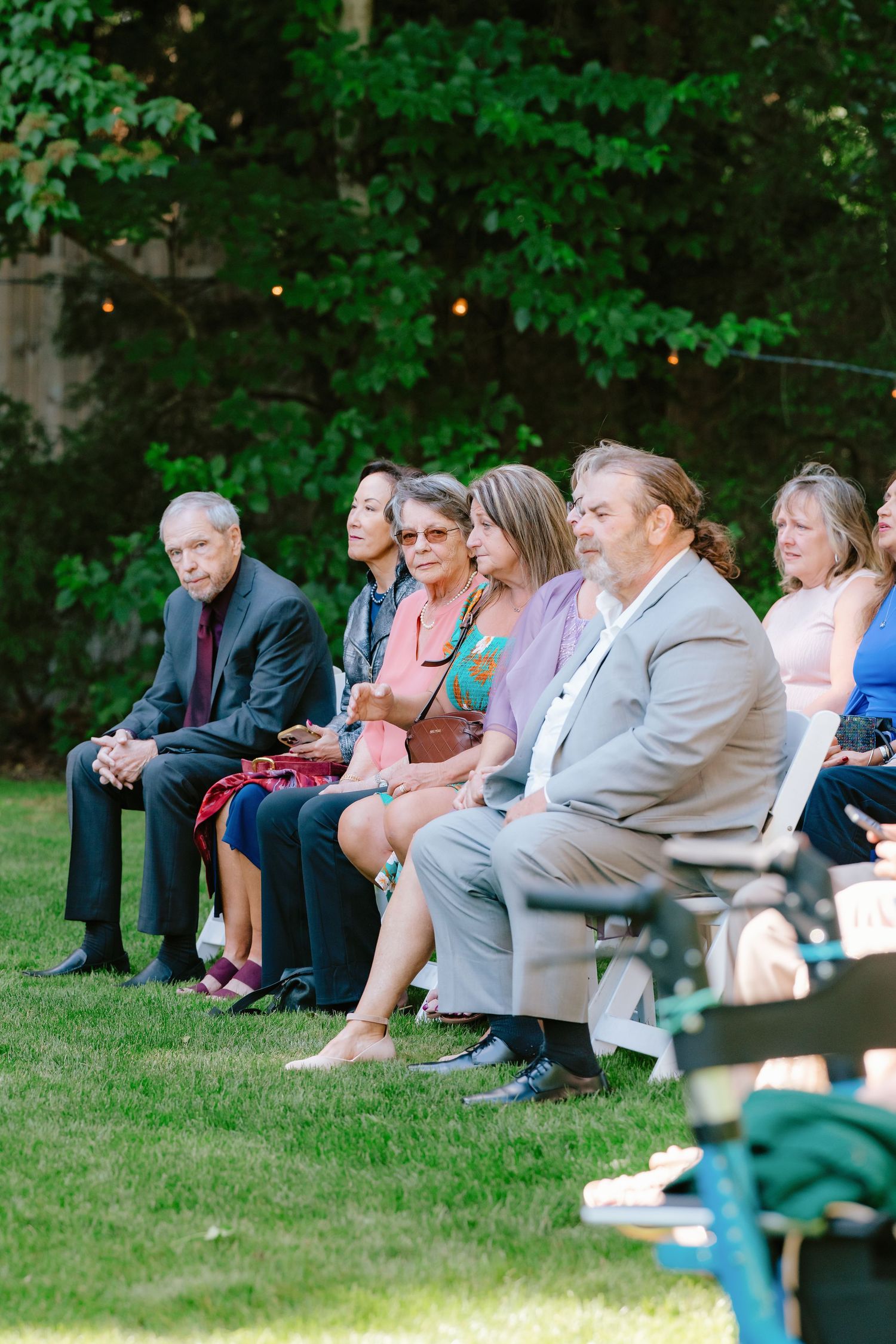 Guests seated in white chairs on grass at an outdoor wedding ceremony surrounded by lush green trees.
