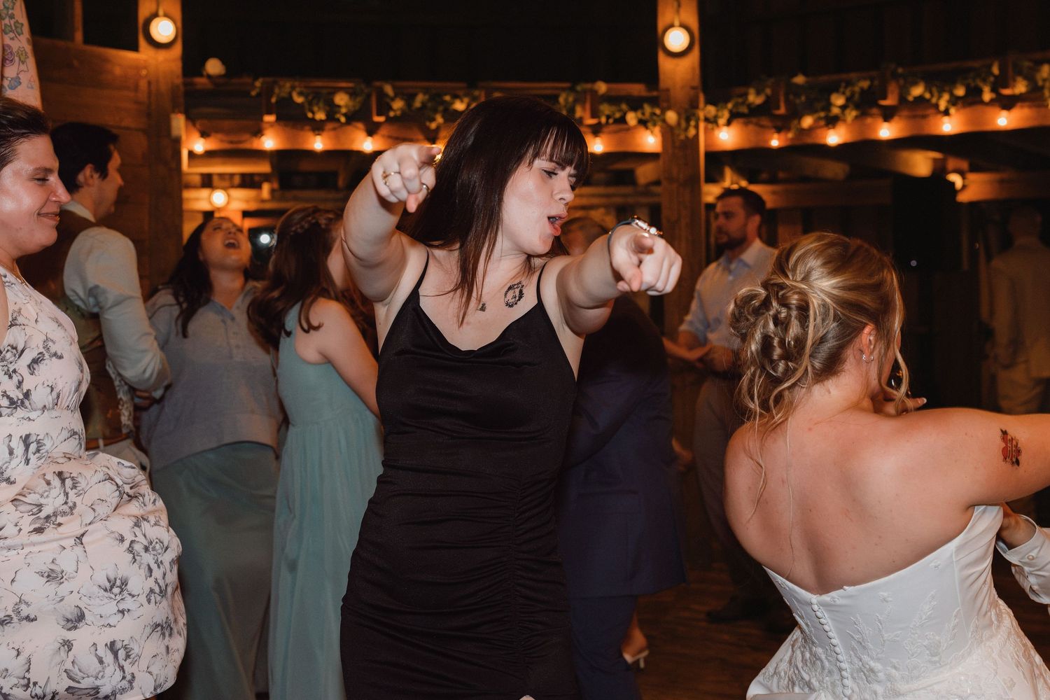 Guests dancing at a rustic wedding reception, woman in black dress pointing while bride in strapless gown dances nearby.