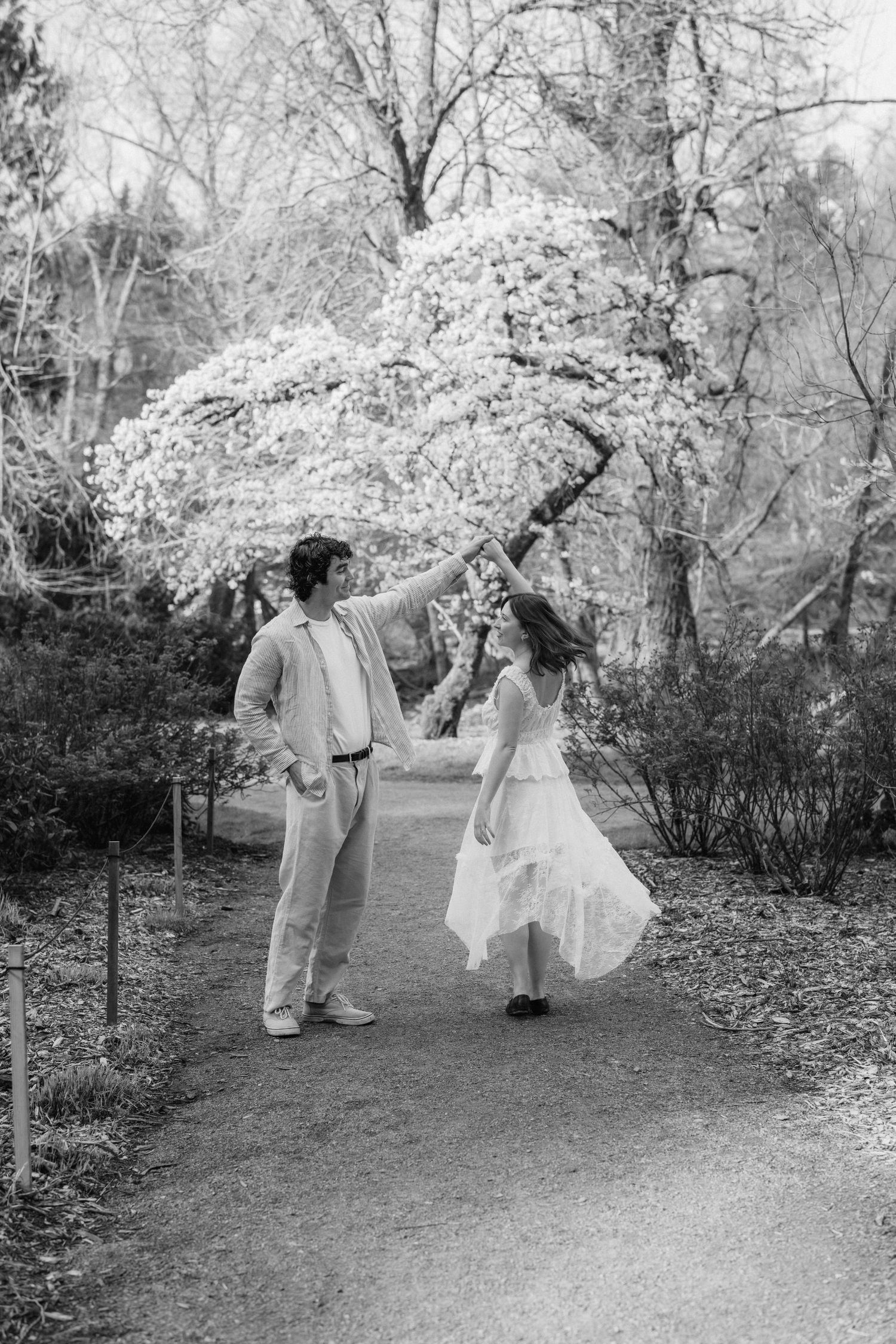 Black and white photo of a couple dancing on a garden path, with blooming trees in the background.