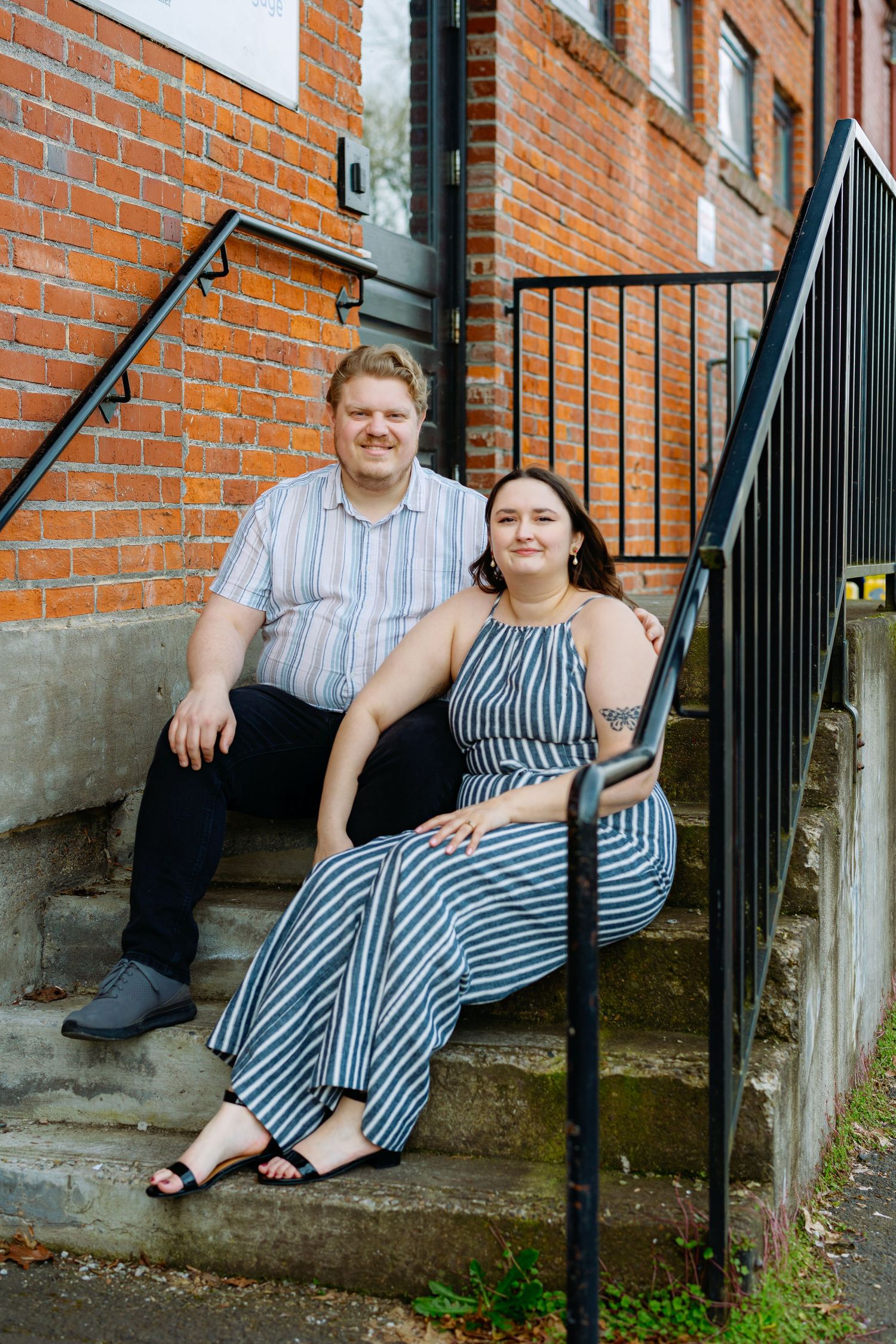 A couple sits together on concrete steps against a brick building with black railings.