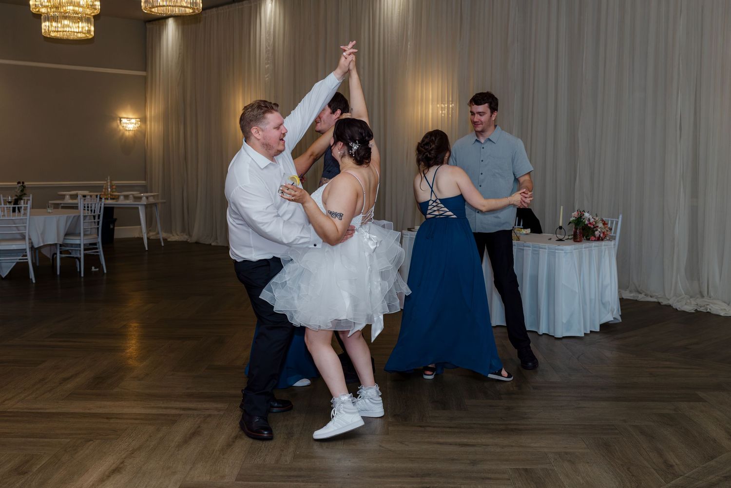 Bride and groom dancing together with wedding party guests on wooden dance floor at reception.