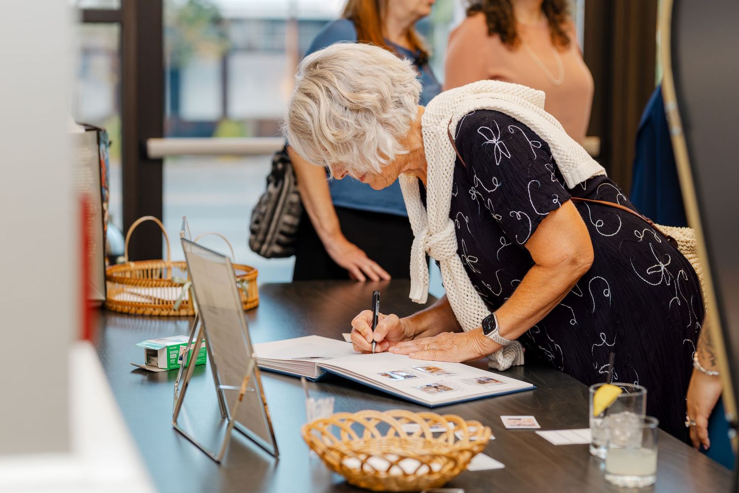 Elderly woman in black floral dress signs wedding guest book at reception table with decorative basket.