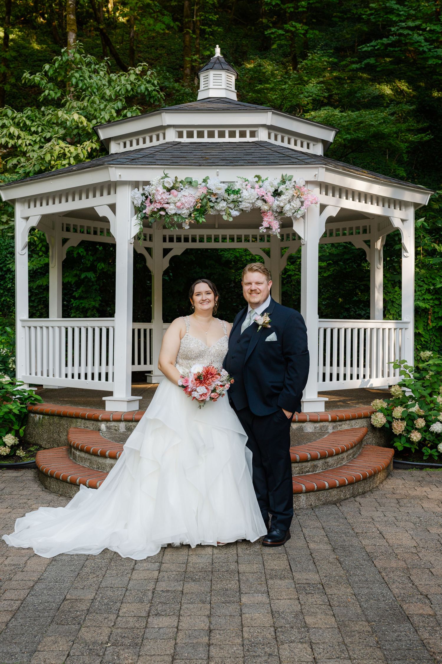 Bride and groom posing in front of a white floral-adorned gazebo on their wedding day.