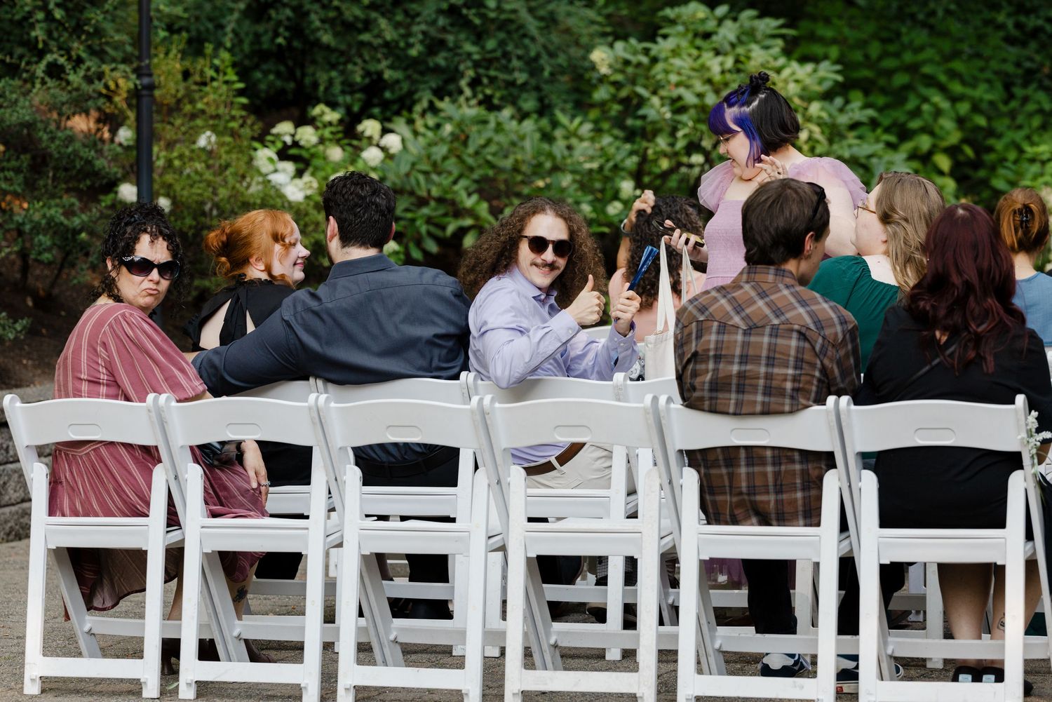 Wedding guests seated in white chairs outdoors, smiling and taking photos during a garden ceremony.