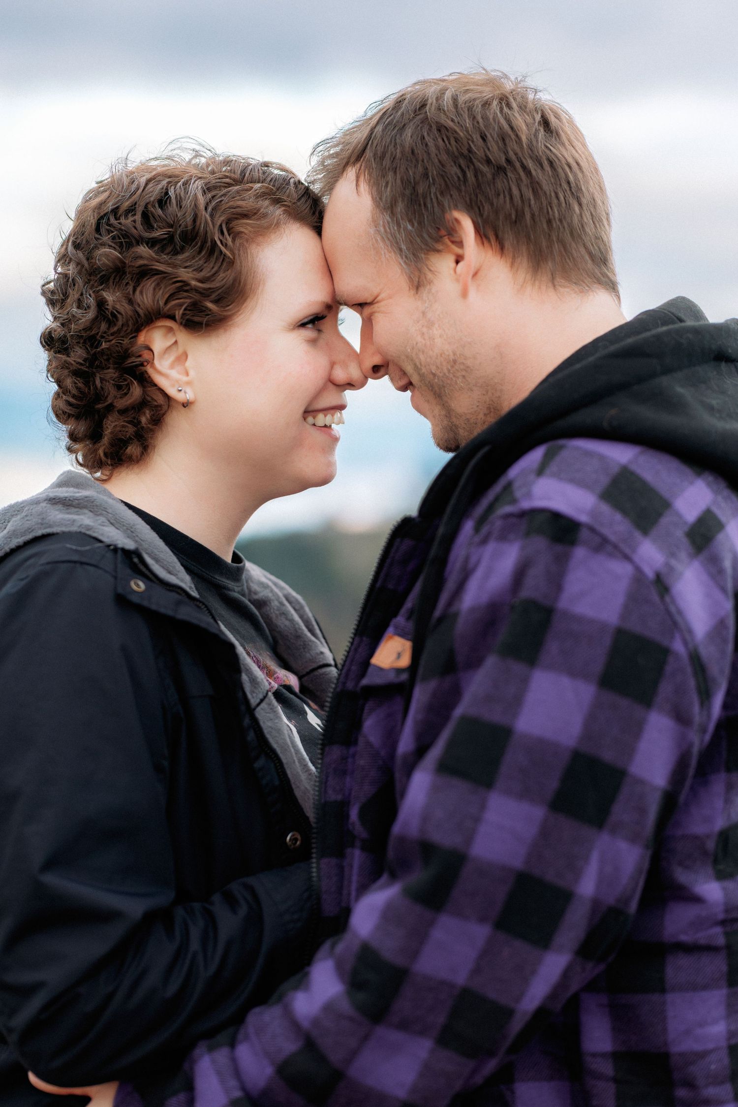 A romantic couple shares intimate moments against a cloudy sky while wearing casual winter clothing.