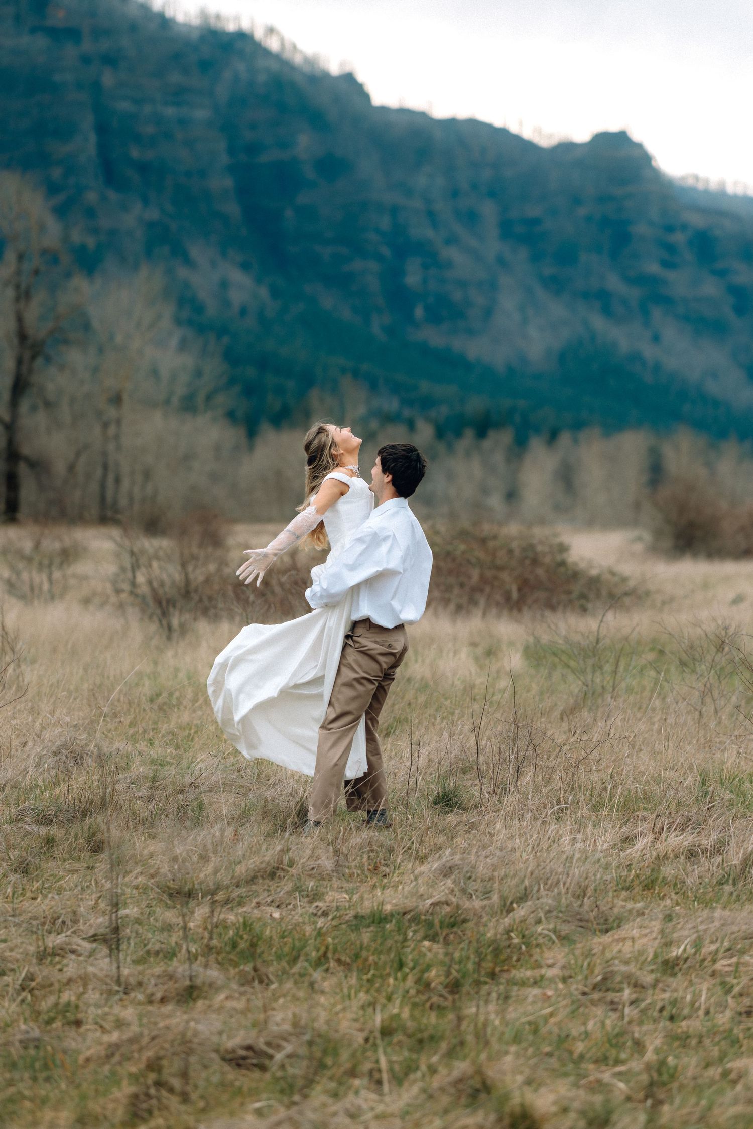 A couple dancing and spinning in a rustic field with mountains in the background on an overcast day.