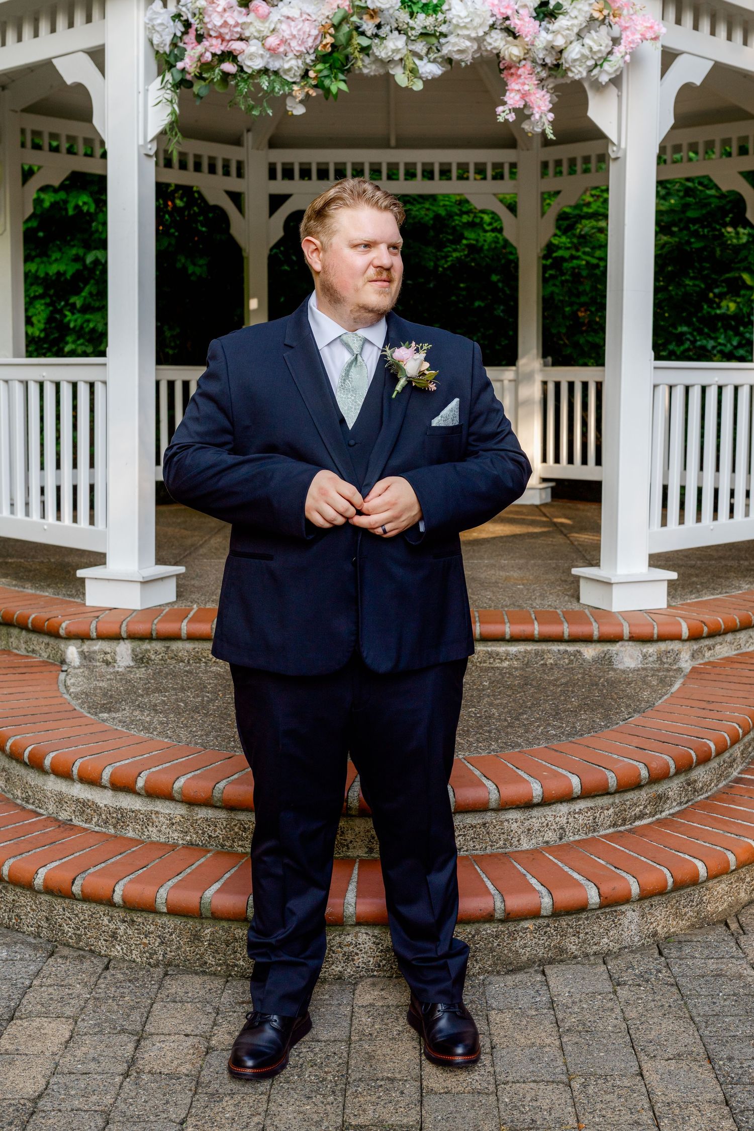 Groom in navy suit with boutonniere stands confidently in front of decorated white wedding gazebo.