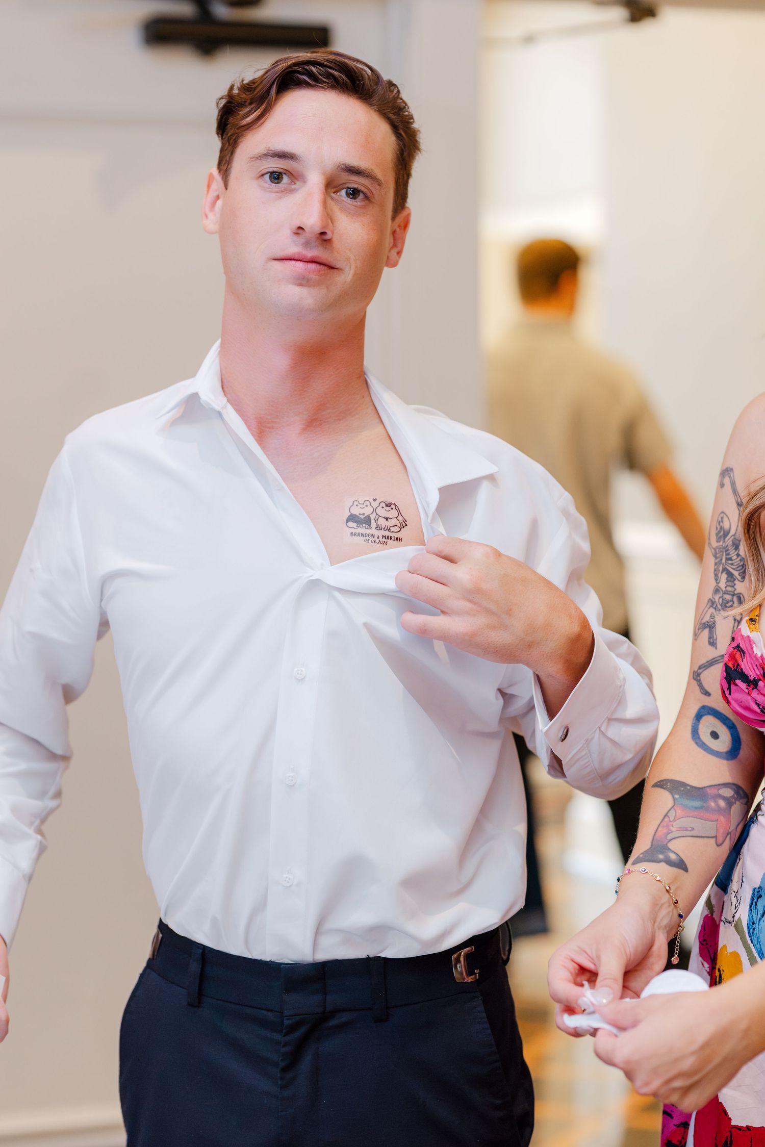 Man in white dress shirt pulling collar open to reveal chest tattoo at a formal event.