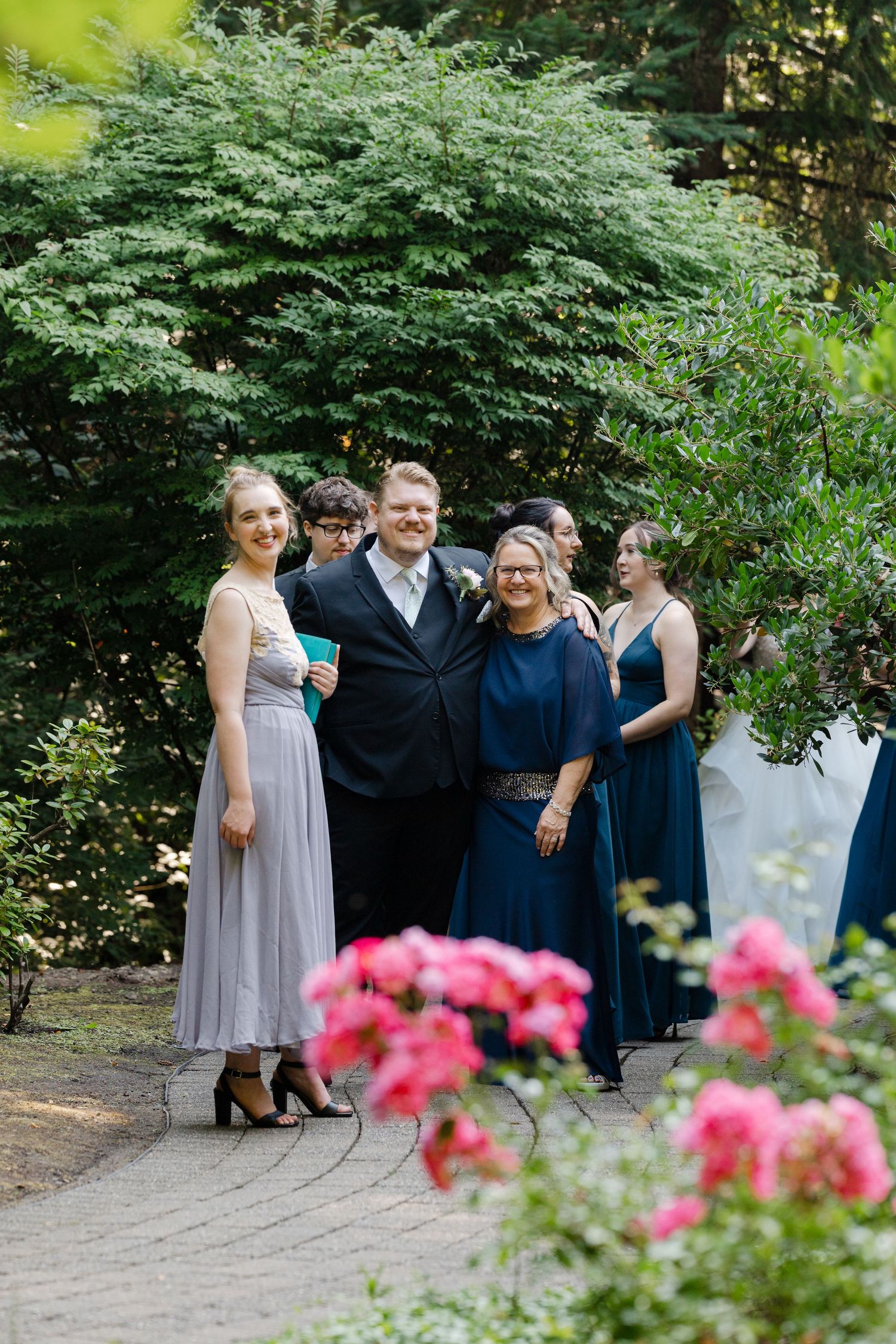 Groom with family members in formal attire posing on garden path with pink roses in foreground.