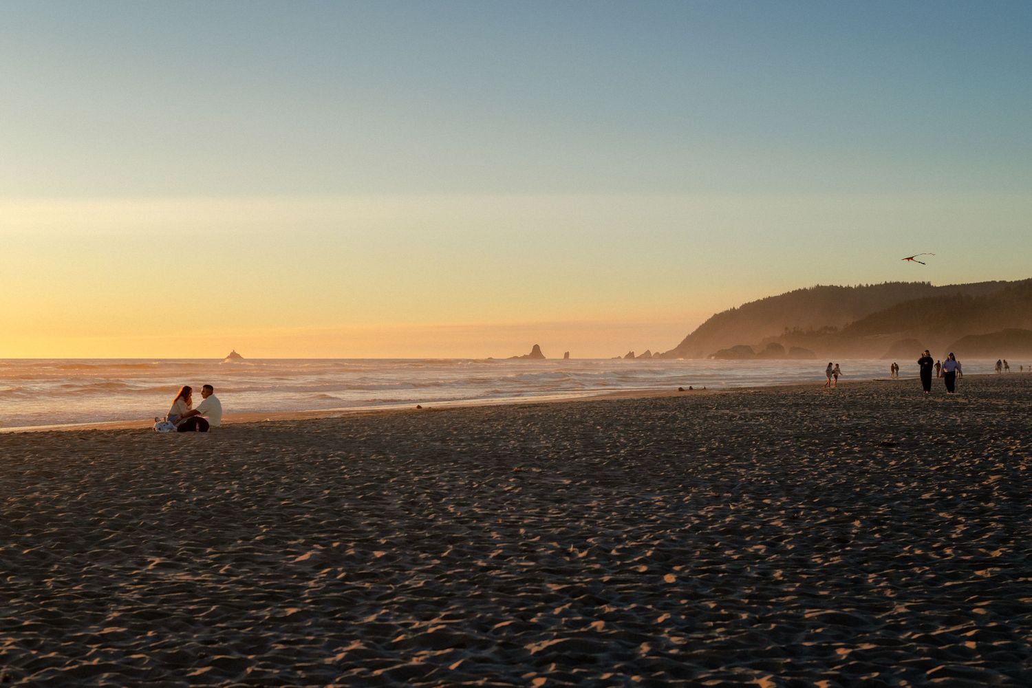 Sunset beach scene with silhouetted people on dark sand, golden sky, ocean waves, and misty coastal cliffs in the distance.