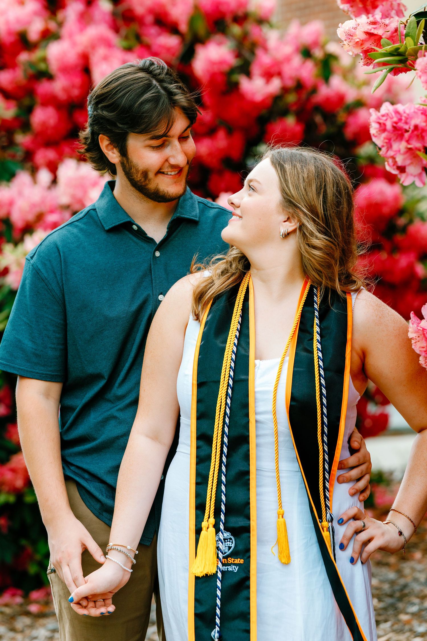 Couple smiling at each other in front of pink flowering bushes, woman wearing graduation stole.