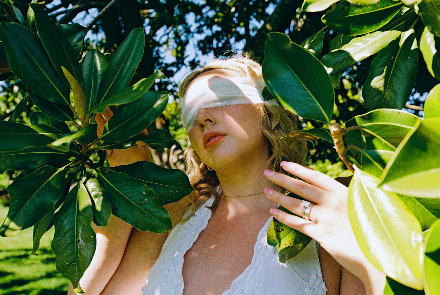 Blonde woman blindfolded with white fabric surrounded by green magnolia leaves in sunlit garden, wearing white dress.