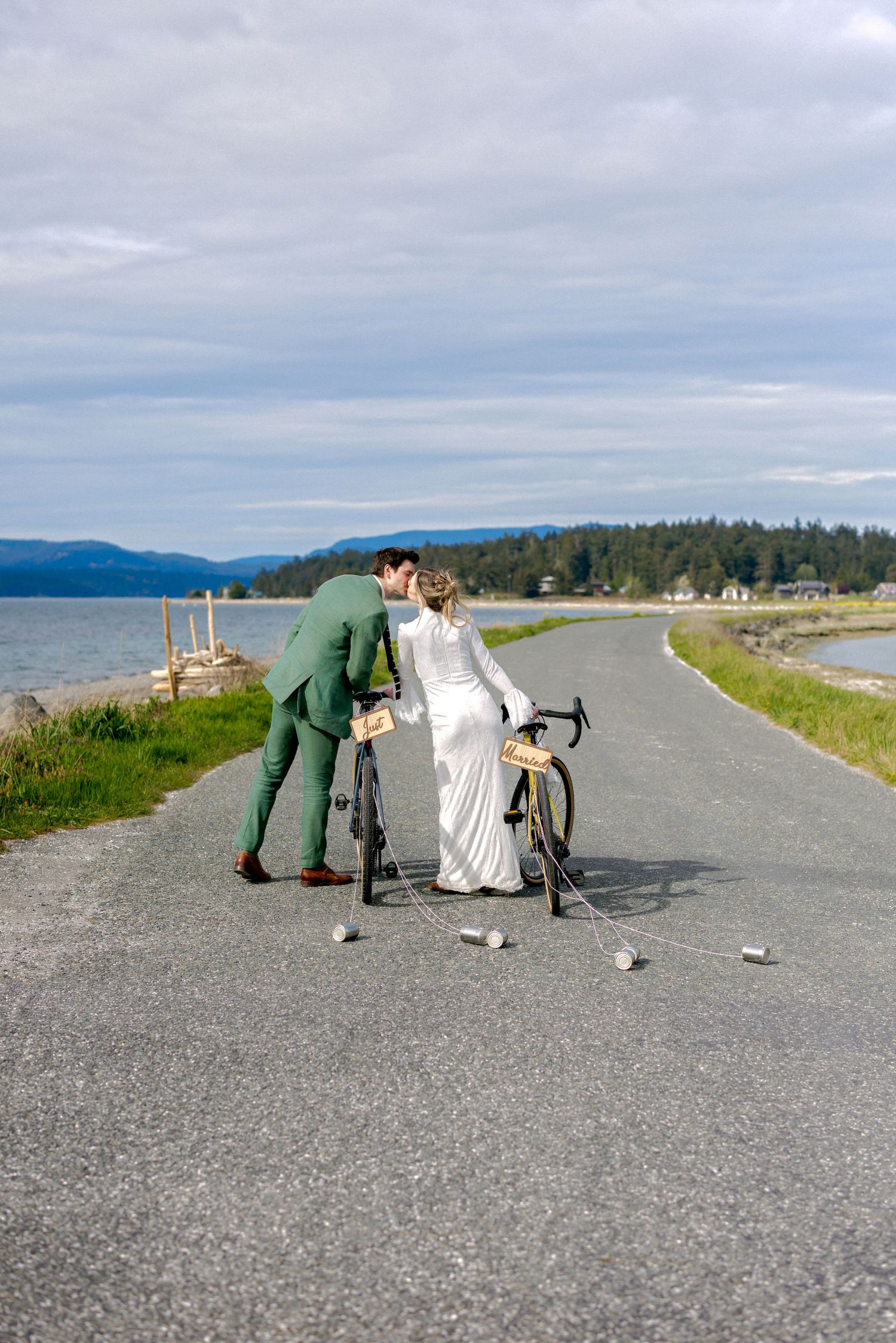 Bride in white gown and groom in green suit kiss over a road bicycle on scenic coastal road.